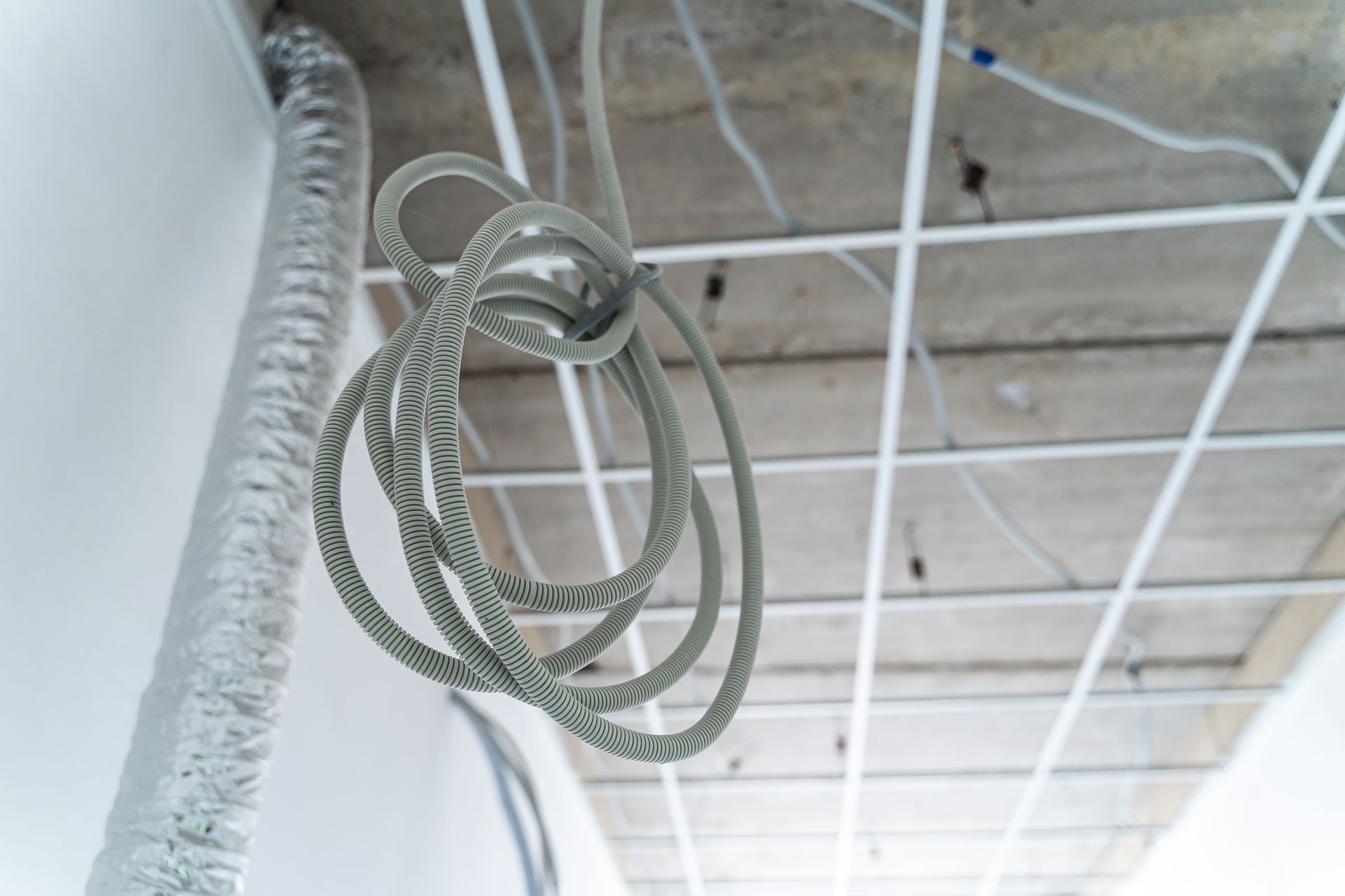 Dans une pièce en cours d'aménagement, une bobine de conduit électrique gris flexible est suspendue à une grille de plafond suspendu.