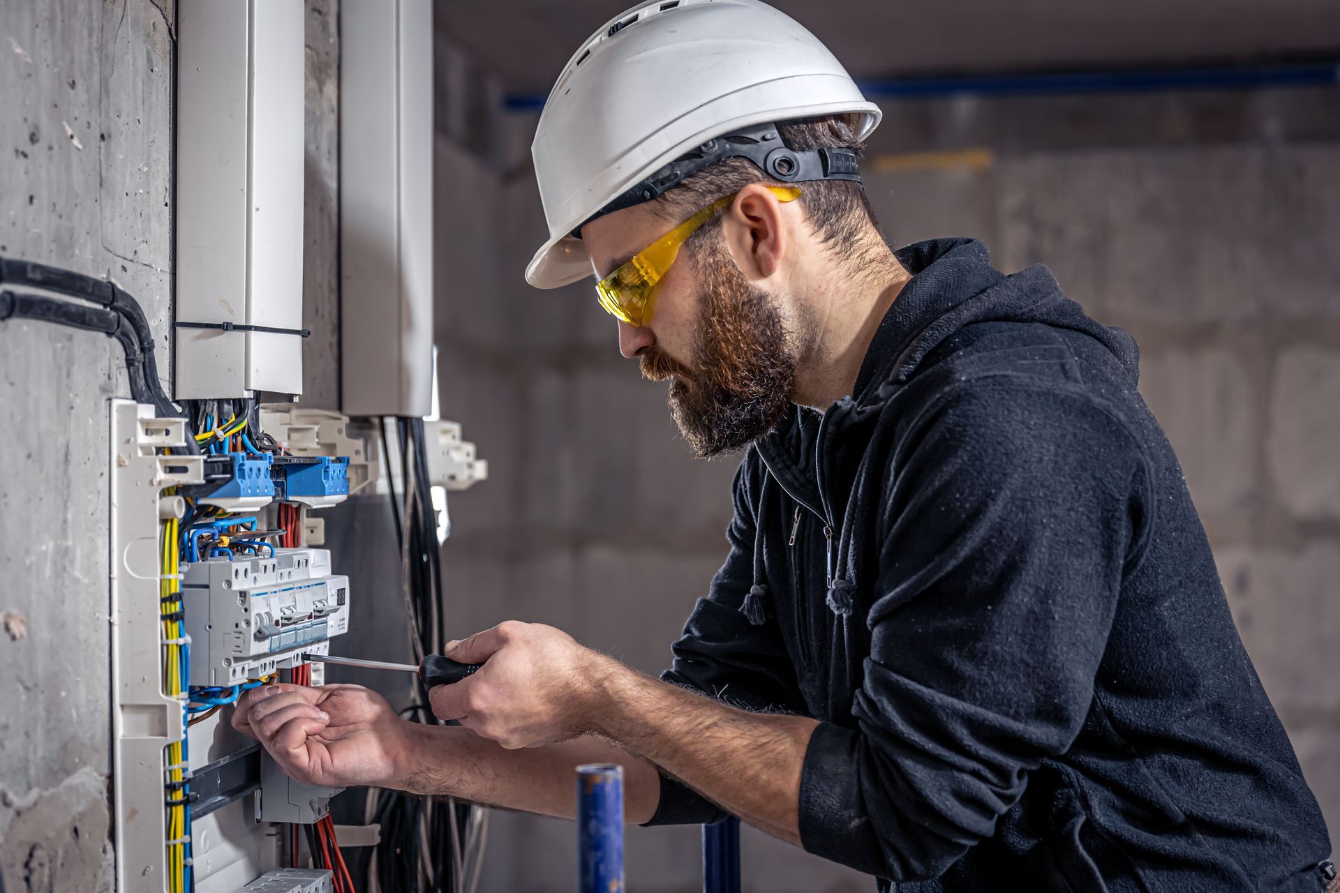 Un électricien professionnel, portant un casque et des lunettes de sécurité, travaille sur un tableau électrique dans une pièce en cours de finition.