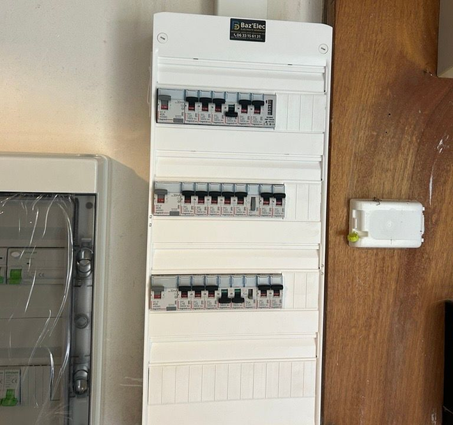 Tableau électrique blanc fixé sur un mur en bois, avec un plus petit tableau gris à côté et un petit appareil blanc à proximité.