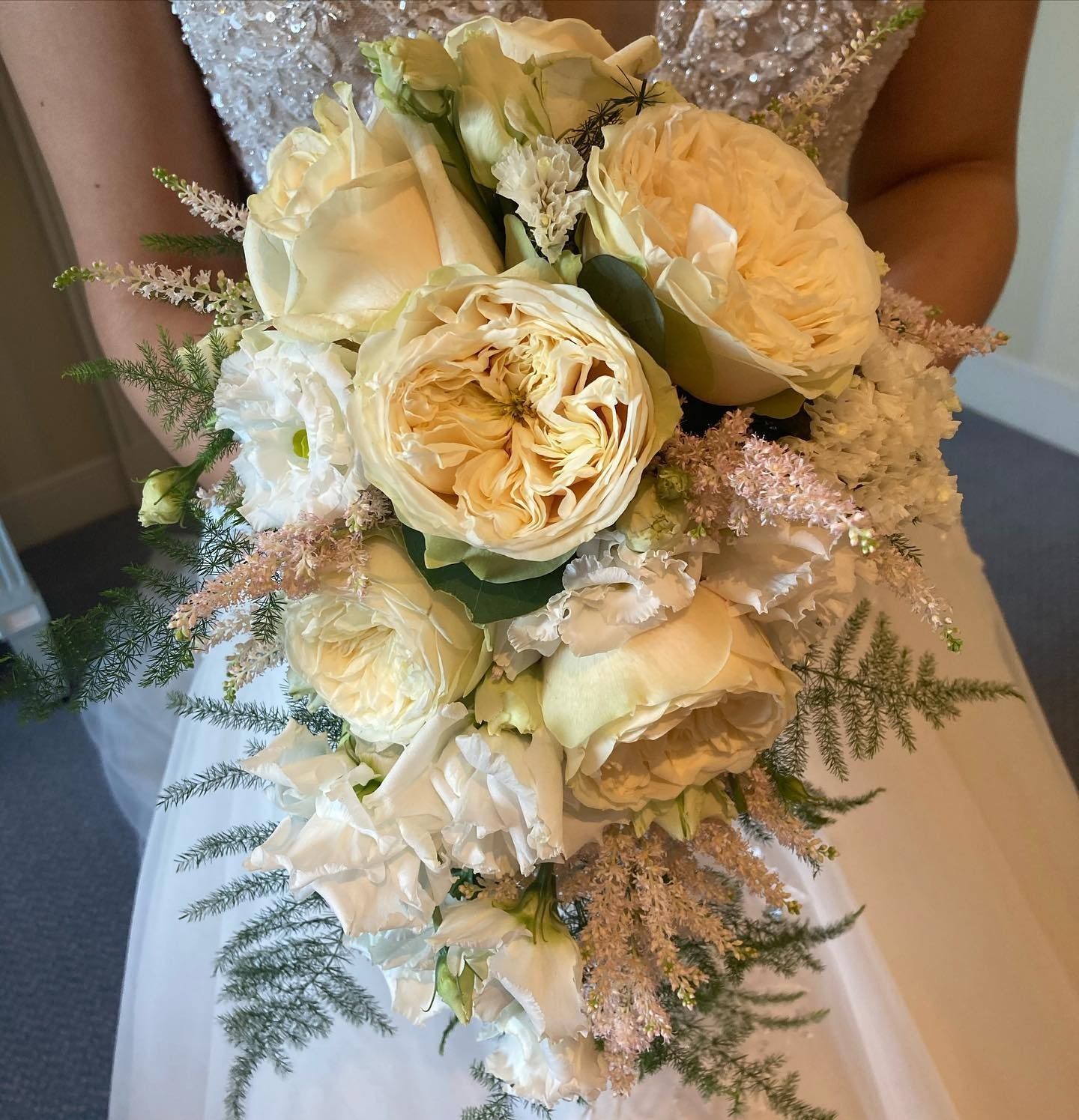 Bouquet de mariée composé de roses blanches et de verdure, tenu devant une robe de mariée scintillante