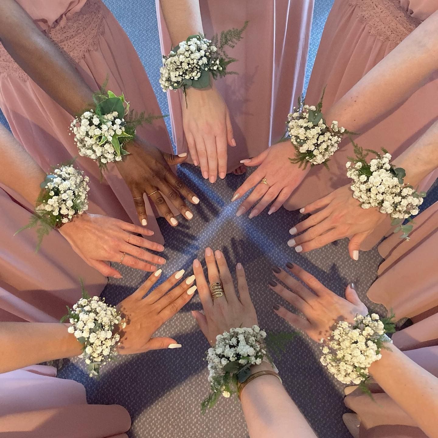 Plusieurs mains disposées en cercle, chacune ornée d'un bracelet de fleurs blanches, sur un tapis bleu.