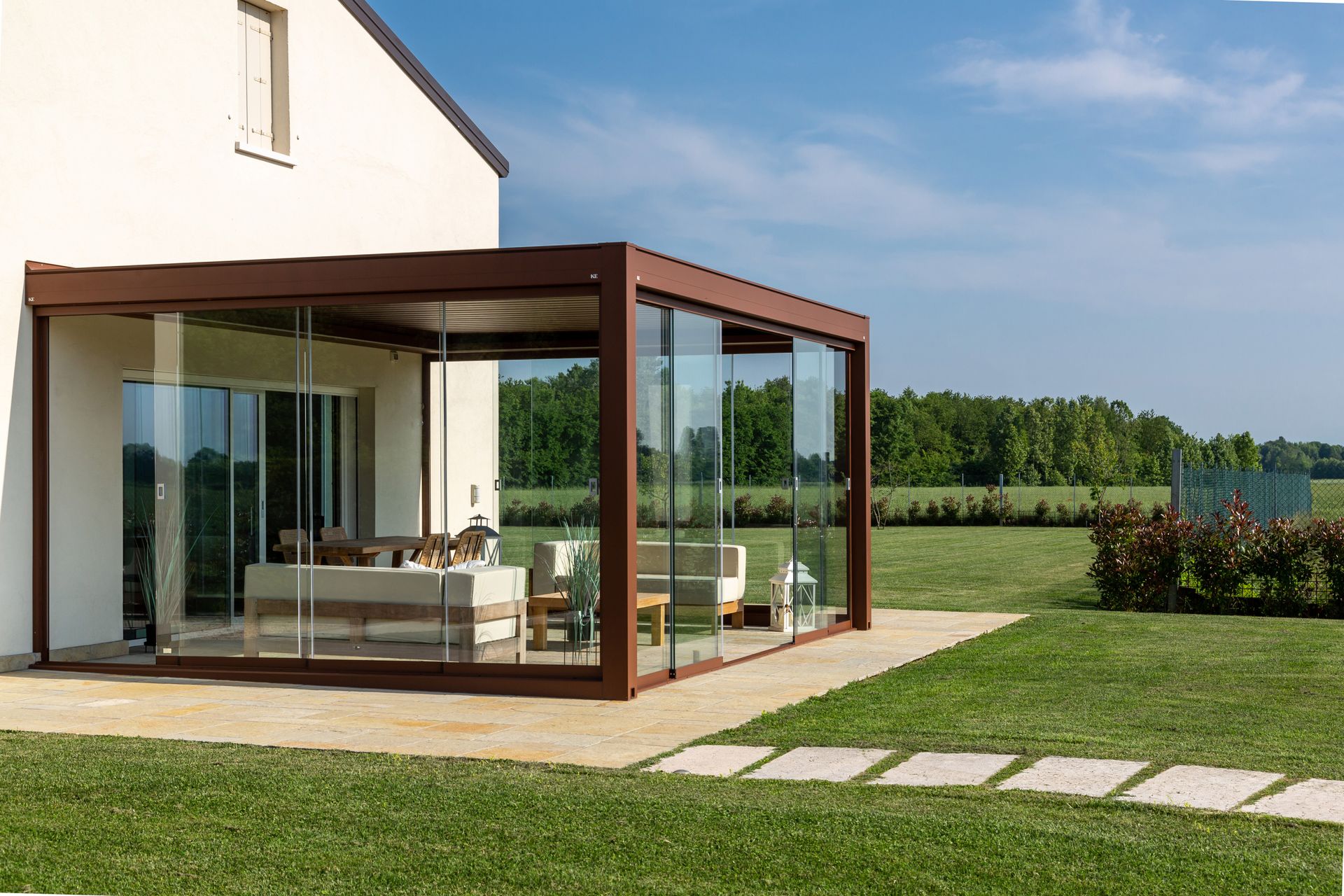 Un moderno patio vetrato annesso a una casa beige, affacciato su un prato e sugli alberi sotto un cielo azzurro e limpido.