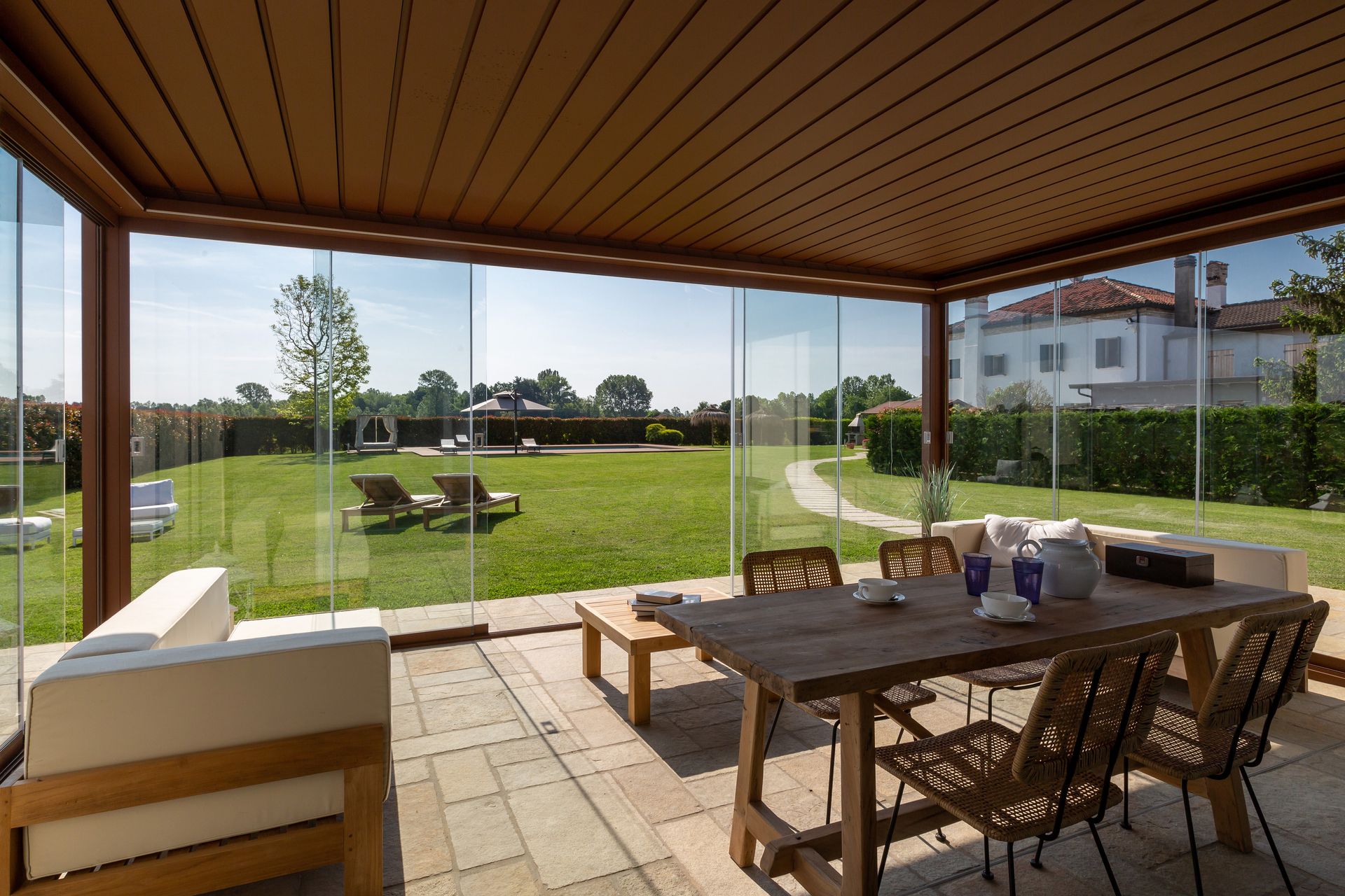 Un patio esterno con set da pranzo e mobili da salotto in legno, racchiuso da pareti di vetro e affacciato su un prato.