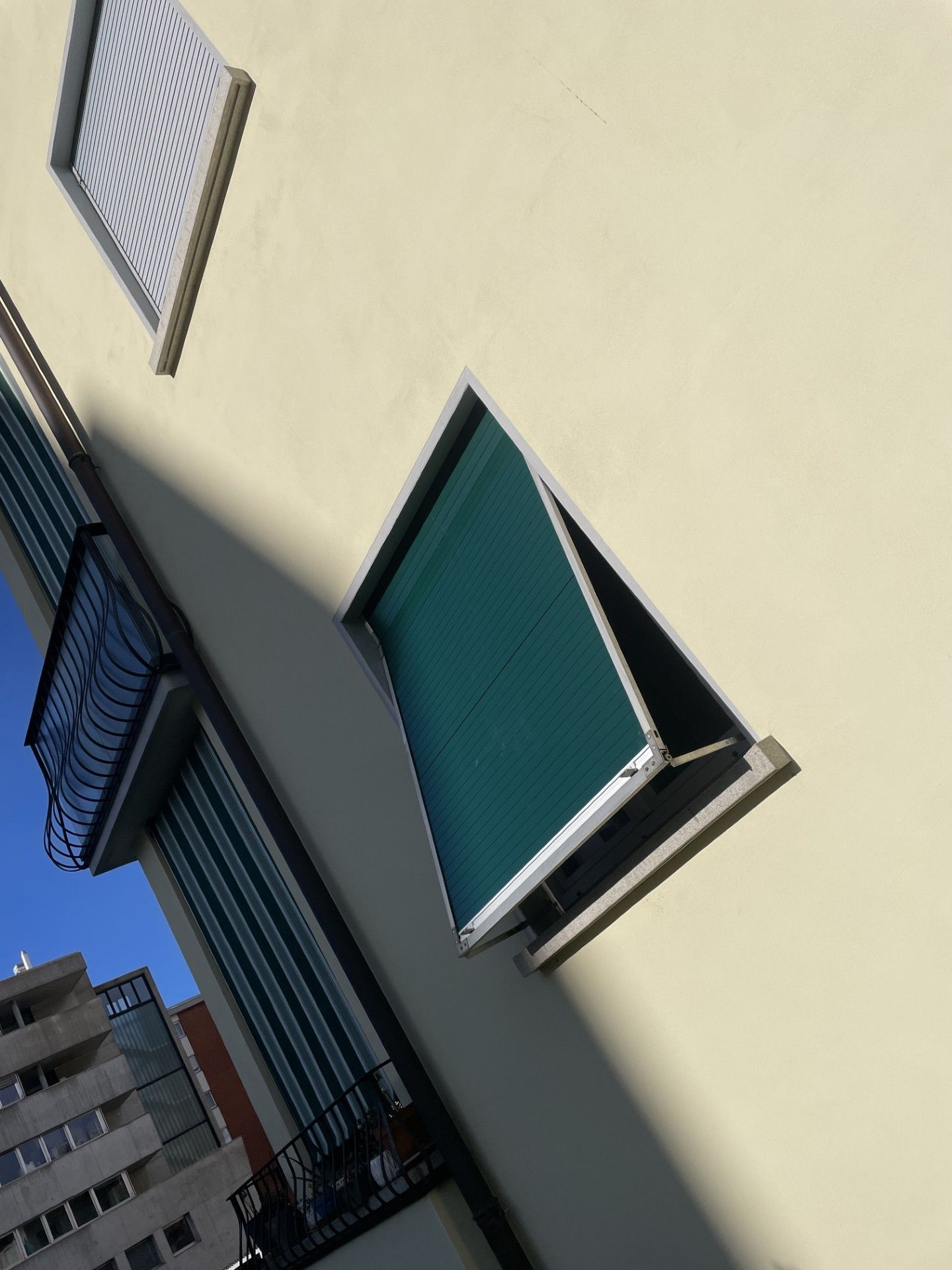 Vista dal basso della facciata di un edificio beige con una persiana verde aperta e un balcone parzialmente visibile.