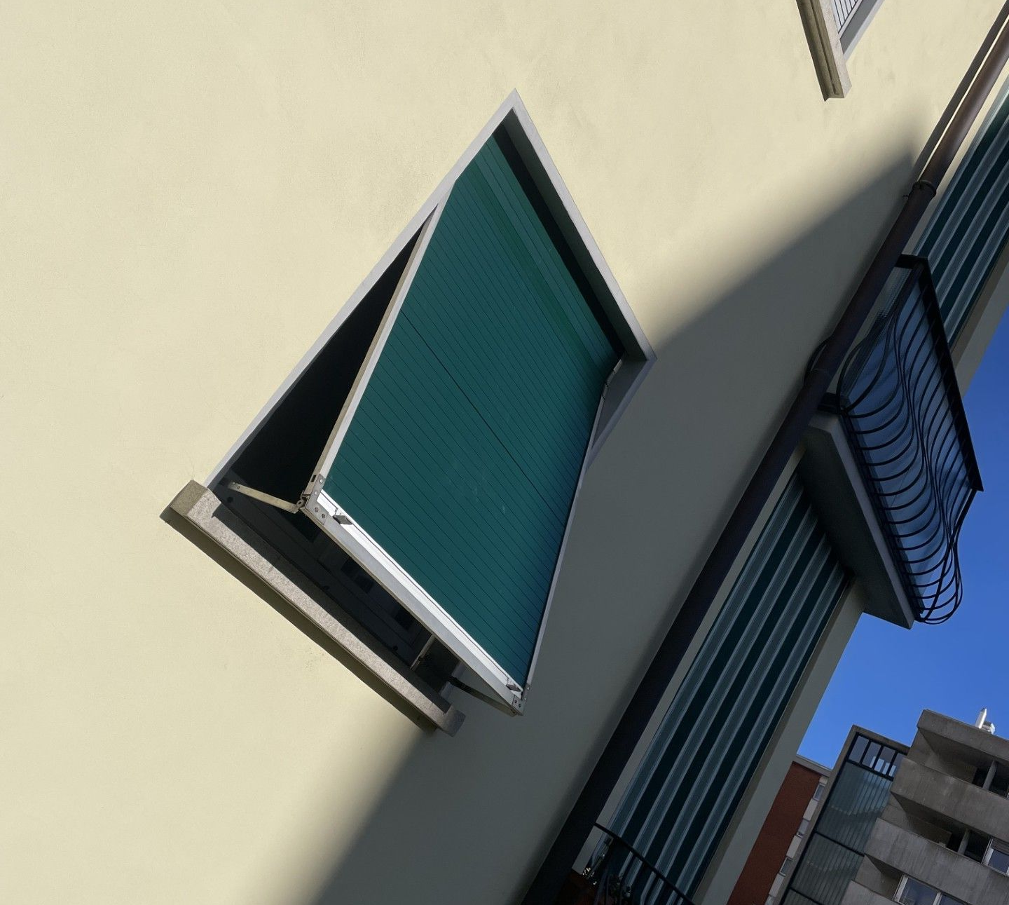 Vista della facciata di un edificio che mostra una parete chiara con una finestra aperta dai vetri verdi e una ringhiera scura del balcone.