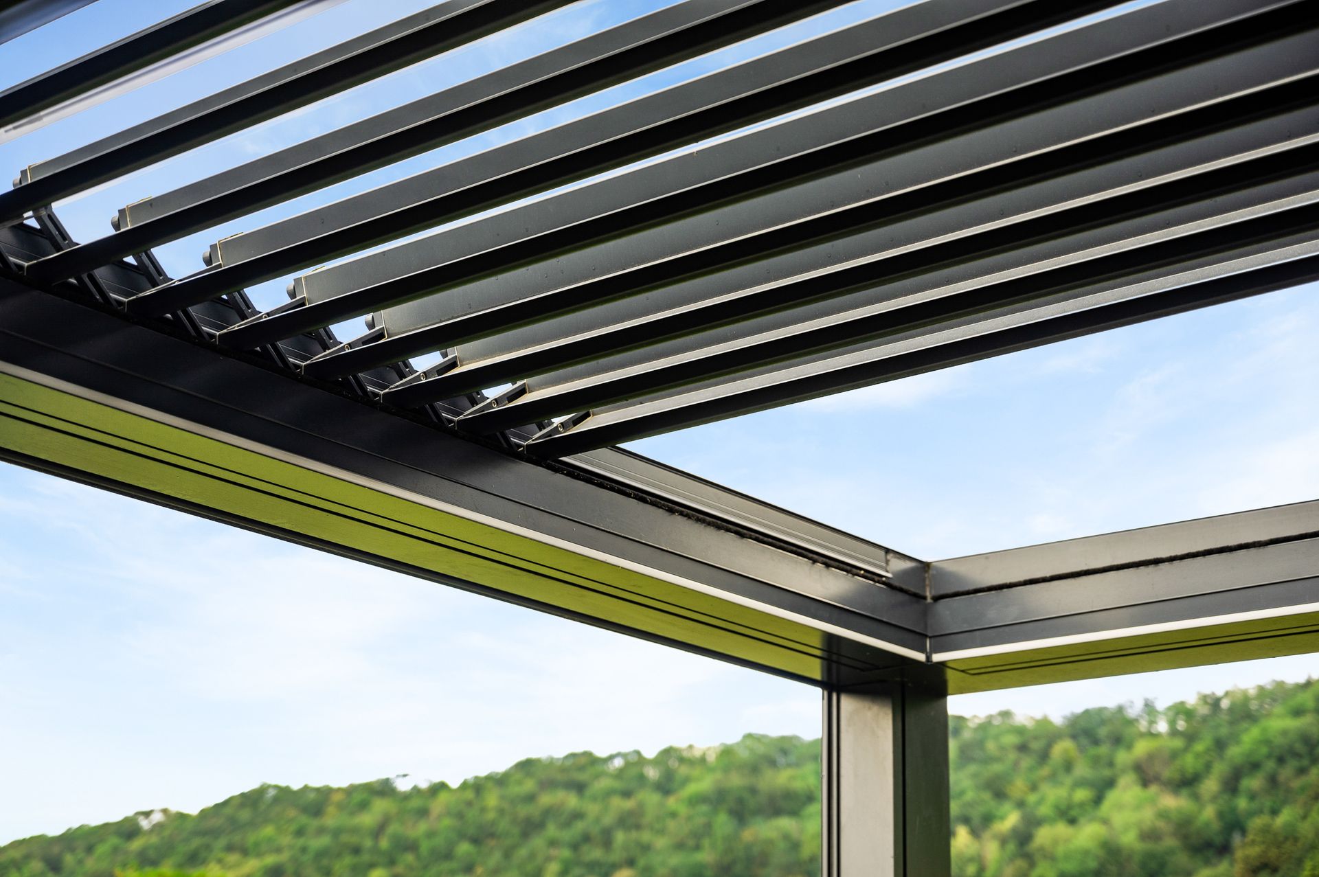 Una vista dal basso di una pergola in metallo scuro con lamelle orientabili, sullo sfondo di un cielo azzurro e di una collina verde.