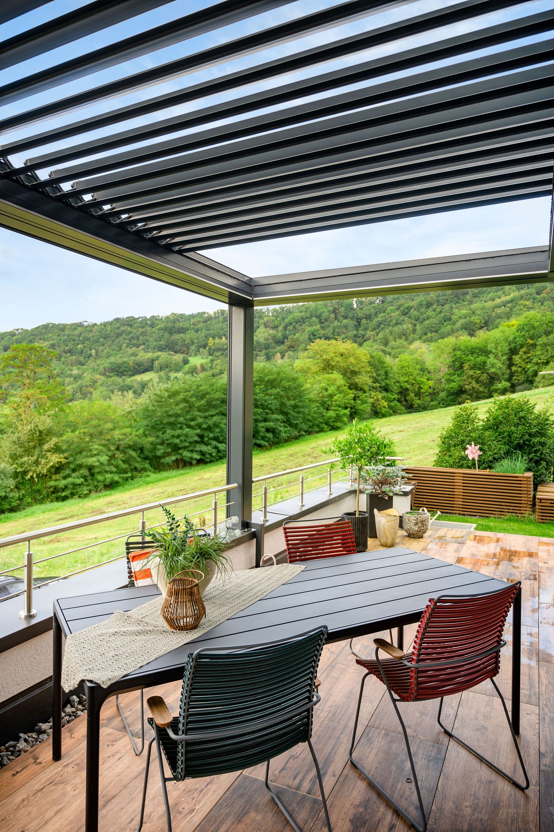 Un moderno patio esterno con tettoia a lamelle, tavolo da pranzo e due sedie intrecciate che si affaccia su un lussureggiante paesaggio collinare verdeggiante.