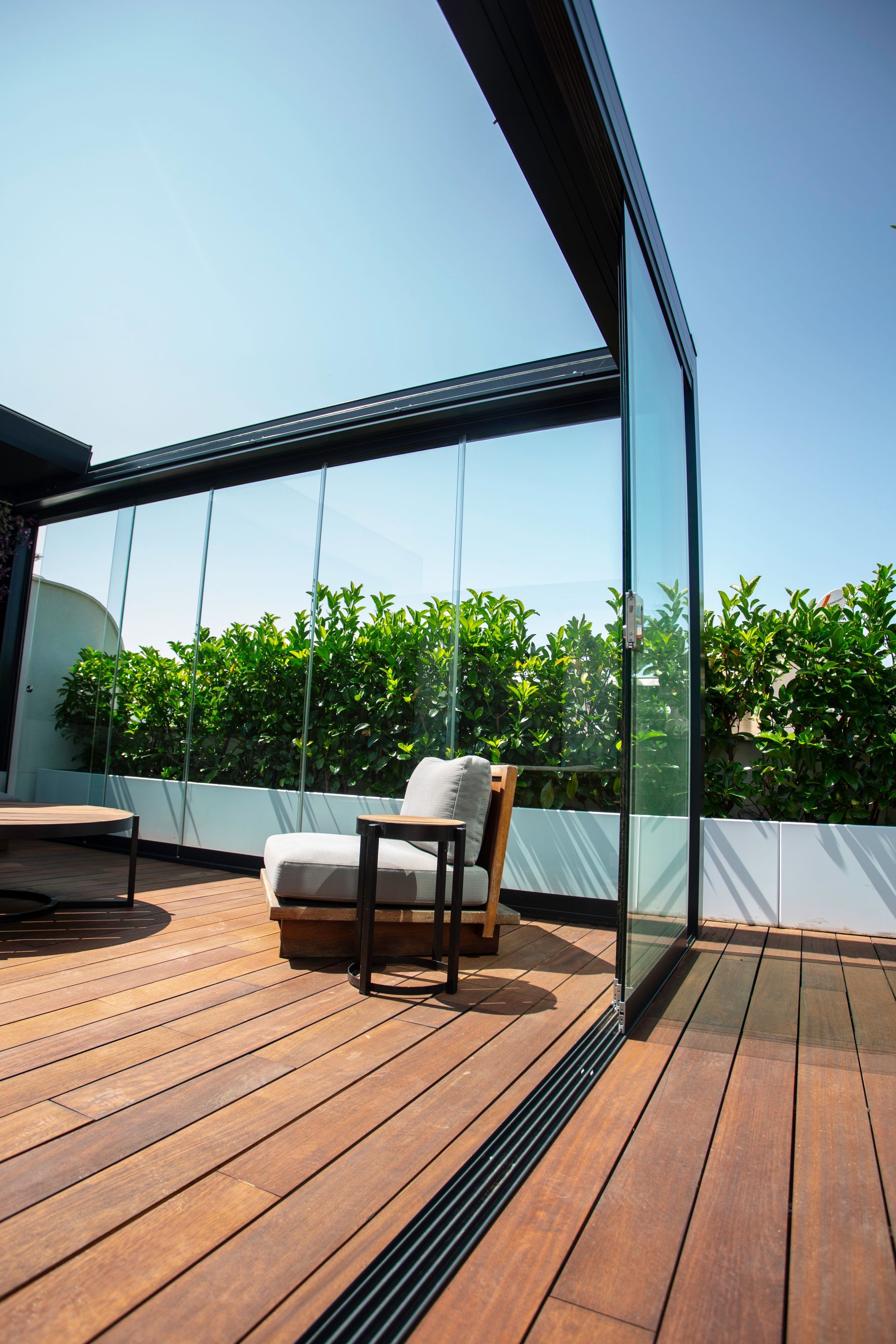 Un patio con una piattaforma in legno, una sedia imbottita con tavolino laterale, pannelli a parete in vetro e una rigogliosa siepe verde.