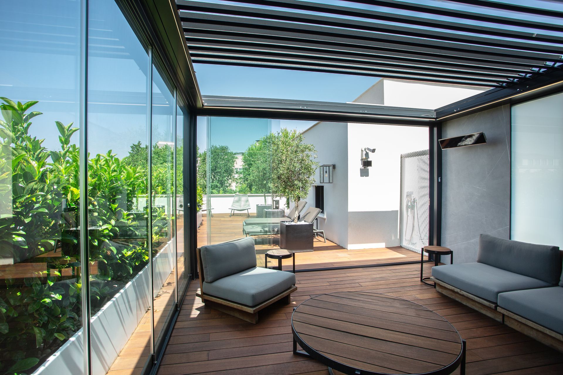 Lounge panoramica moderna e all'aperto con pareti in vetro, pavimento in legno, divani grigi e pergolato con tetto a doghe.