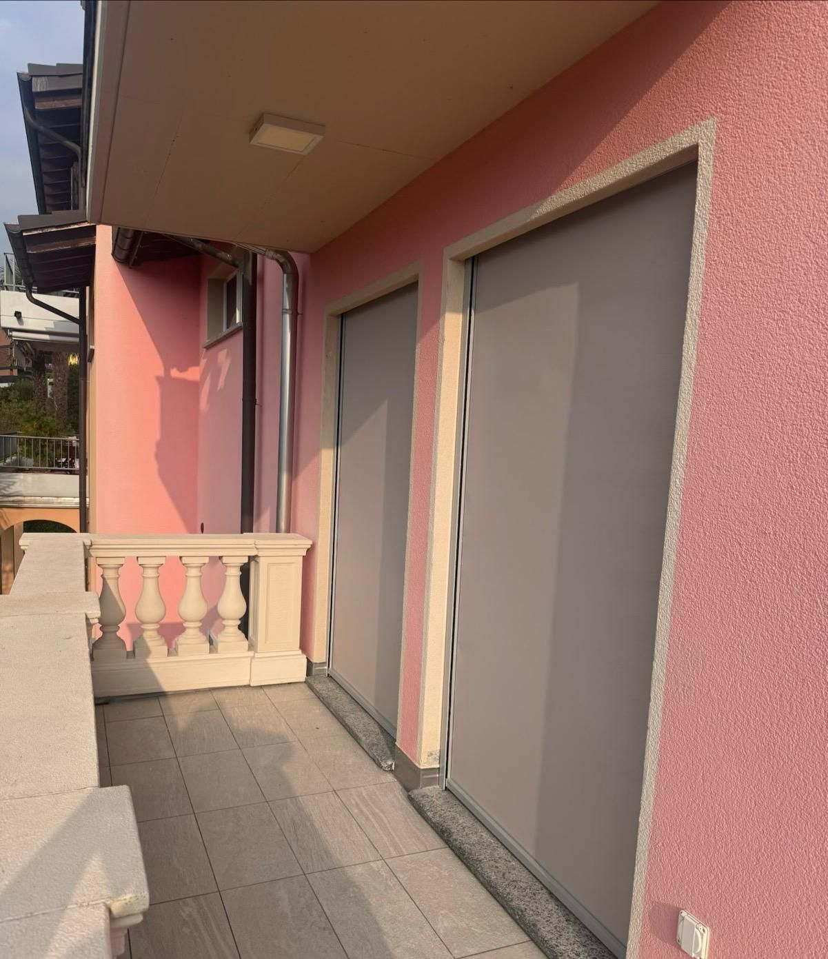Un balcone con pareti rosa dalla texture particolare e due alte porte grigie chiuse, affiancate da una balaustra in pietra chiara.