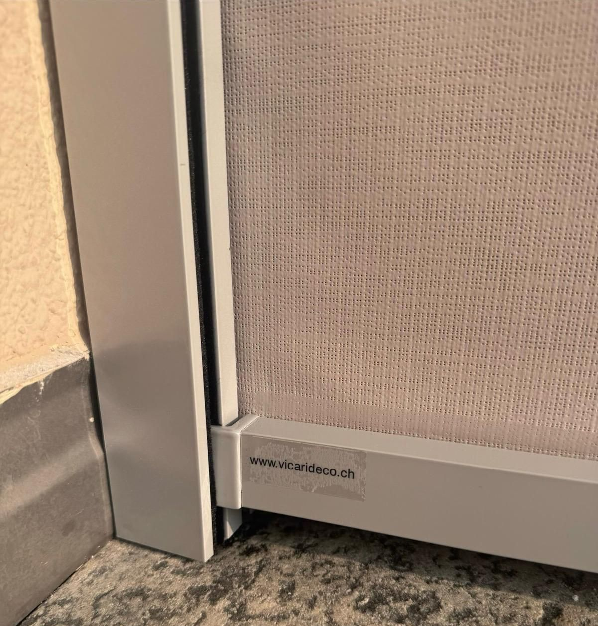 Telaio grigio chiaro e schermo testurizzato di una porta scorrevole a zanzariera installata su un pavimento in pietra.