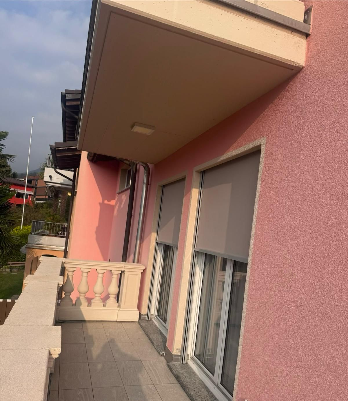 Un balcone con ringhiera in pietra chiara e pavimento in piastrelle, annesso a una casa con pareti dipinte di rosa e con una finitura a rilievo.