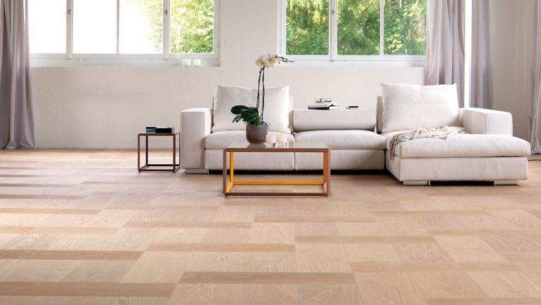 Un pavimento in parquet di legno chiaro conduce a un divano componibile color crema, a un tavolino con una pianta e a grandi finestre.