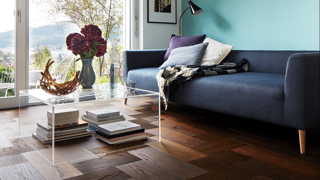 Un soggiorno moderno con divano blu navy, parete turchese, pavimento in piastrelle effetto legno e tavolino da caffè in vetro trasparente con sopra dei libri.