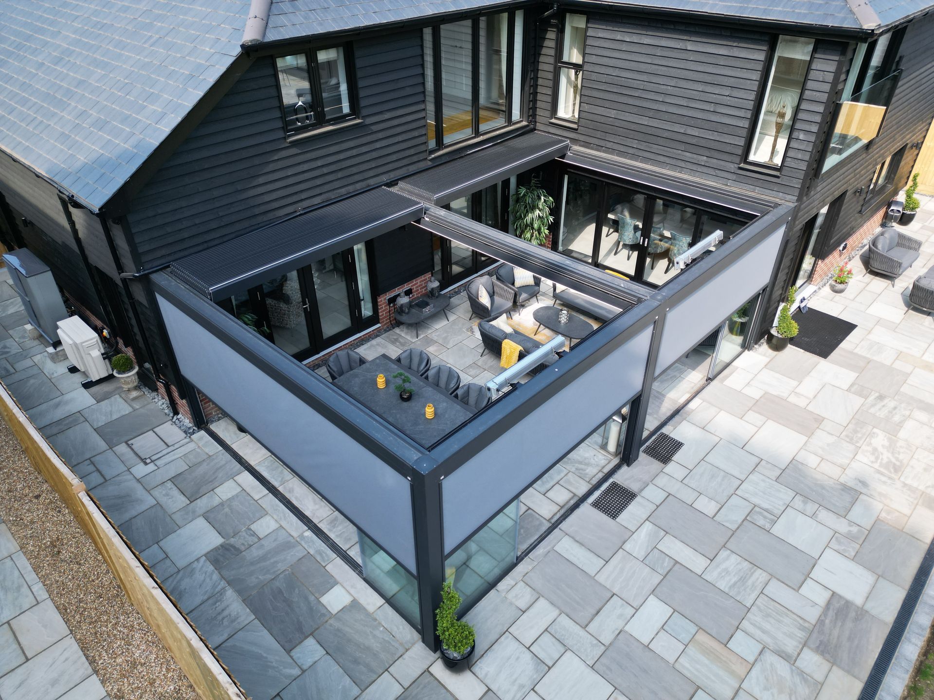 Un patio esterno con una moderna pergola coperta con struttura scura e tende retrattili, annessa a una casa scura.