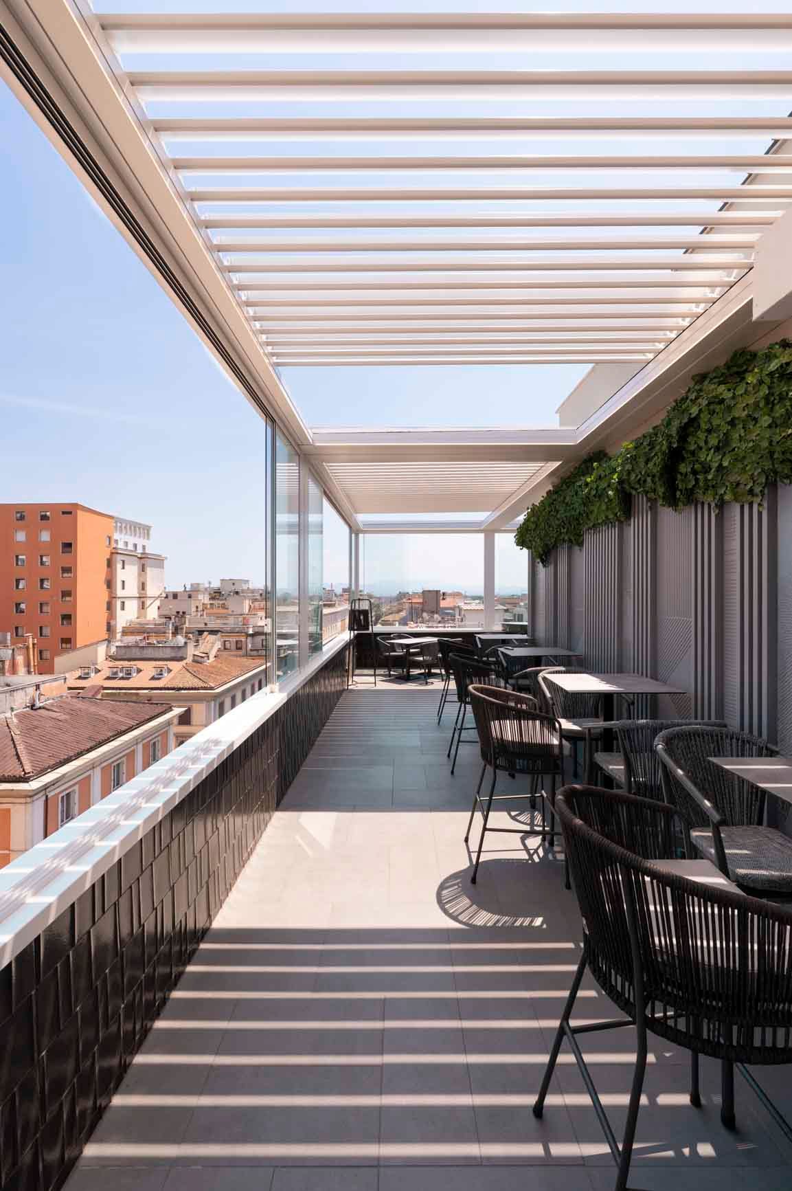 Una soleggiata terrazza ristorante all'aperto con tetto a lamelle retrattili, sedie nere e vista sulla città.