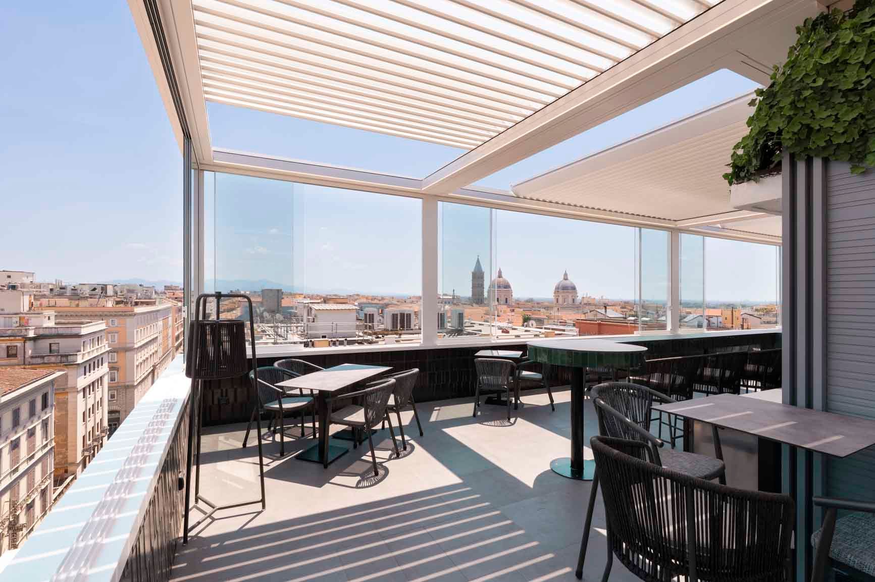 Una terrazza sul tetto con moderni tavoli e sedie in metallo, sotto un tetto a doghe, con vista sullo skyline della città.