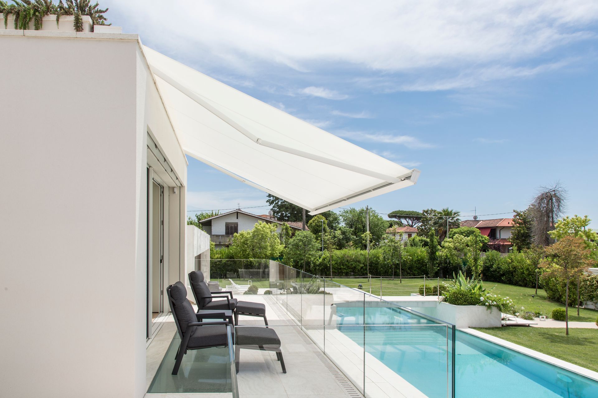 Un patio moderno e bianco con sedie a sdraio e una tenda da sole estensibile che si affaccia su una piscina in una giornata di sole.