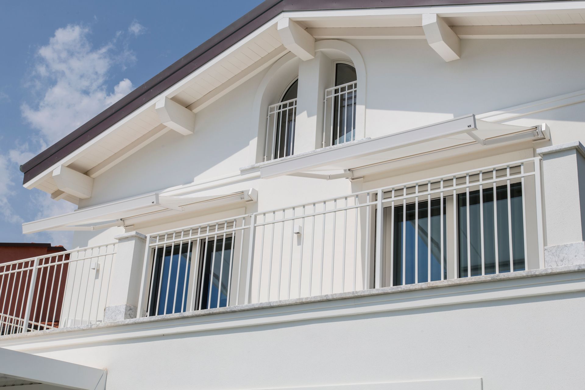 Casa bianca a due piani con balcone, ringhiere in metallo e finestre ad arco sotto un cielo azzurro.
