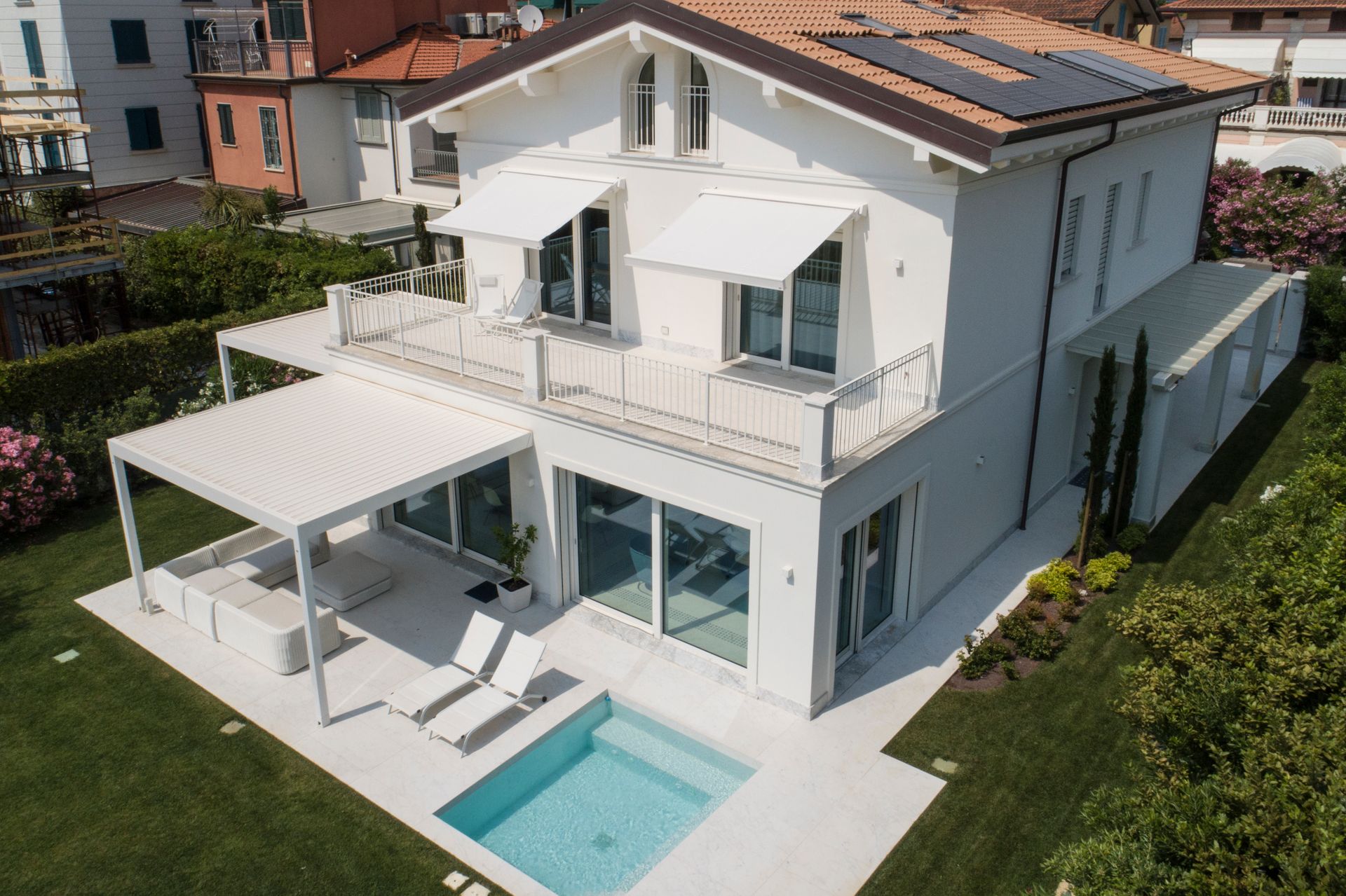 Una villa moderna bianca a due piani con una piccola piscina, mobili da giardino e un pergolato in un soleggiato cortile verde.