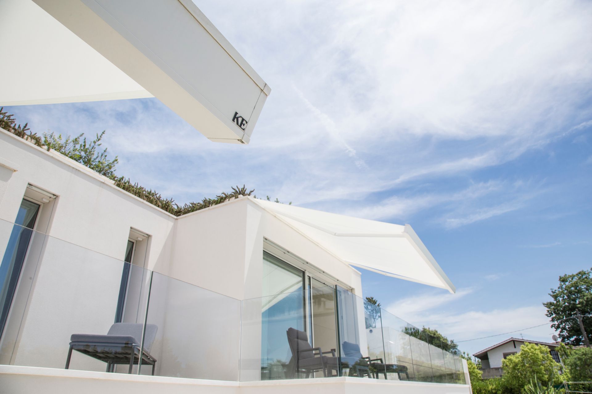 Casa moderna bianca con un'elegante tenda da sole retrattile sopra un patio con ringhiera in vetro, sotto un cielo azzurro brillante.