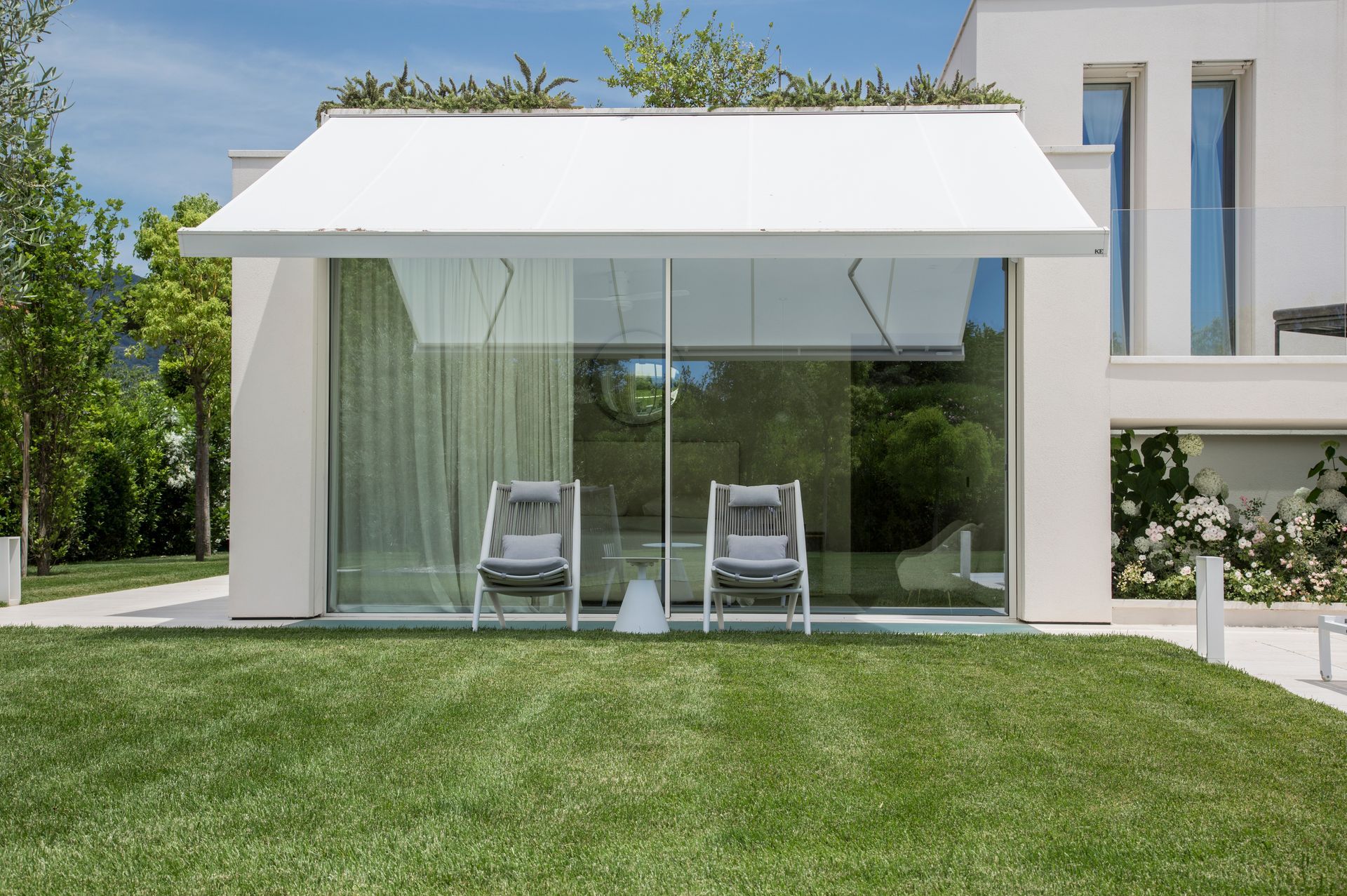 Due sedie da esterno sono posizionate su un prato di fronte a una moderna casa bianca con un'ampia porta a vetri e una tenda da sole bianca retrattile.