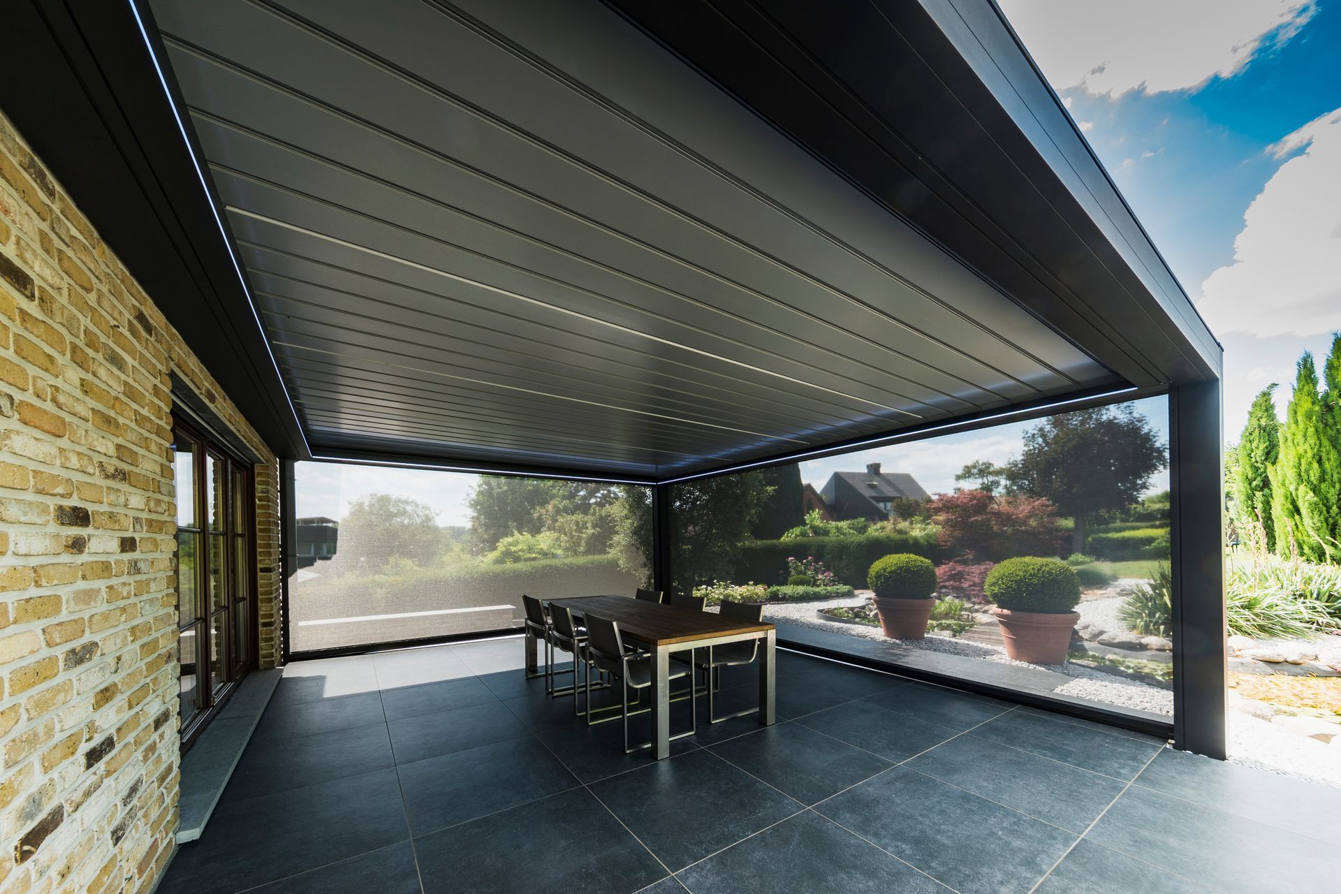Un patio esterno coperto con un elegante tetto nero e zanzariere a scomparsa, arredato con un tavolo da pranzo su un pavimento piastrellato.