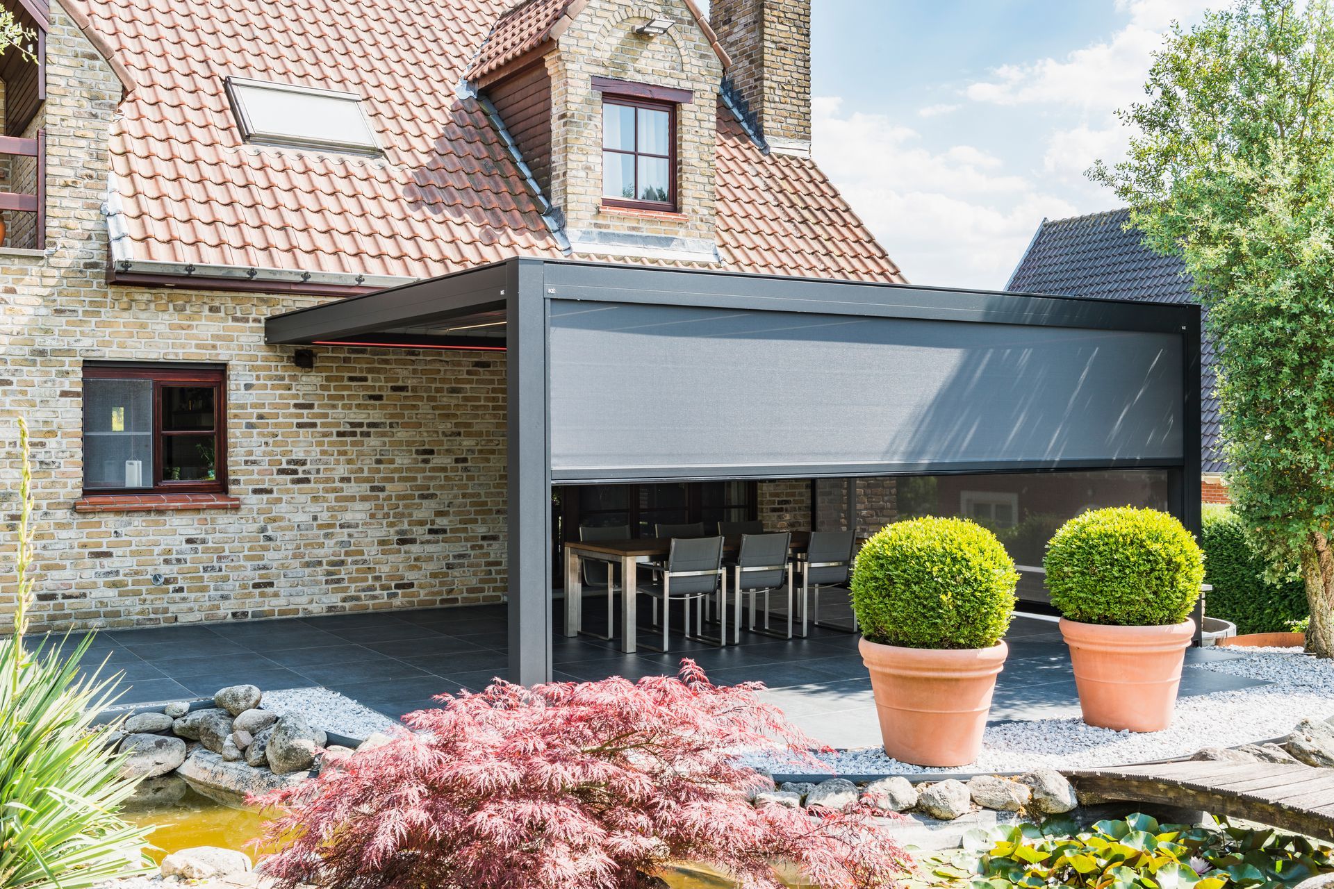 Una casa in pietra con un moderno pergolato in metallo scuro dotato di tenda retrattile e set da pranzo su una terrazza in pietra.