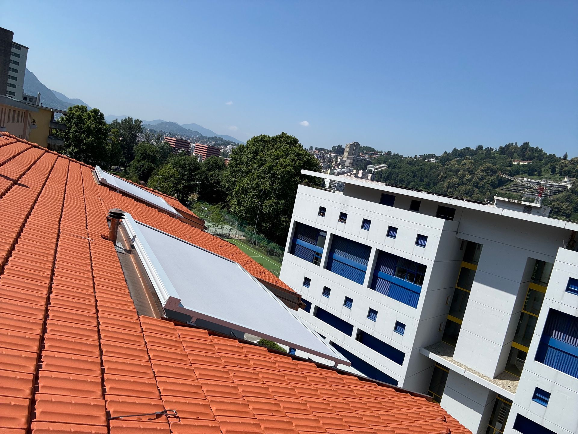 Un tetto di tegole rosse con due lucernari si affaccia su un edificio bianco a più piani, sullo sfondo di alberi e un cielo azzurro.