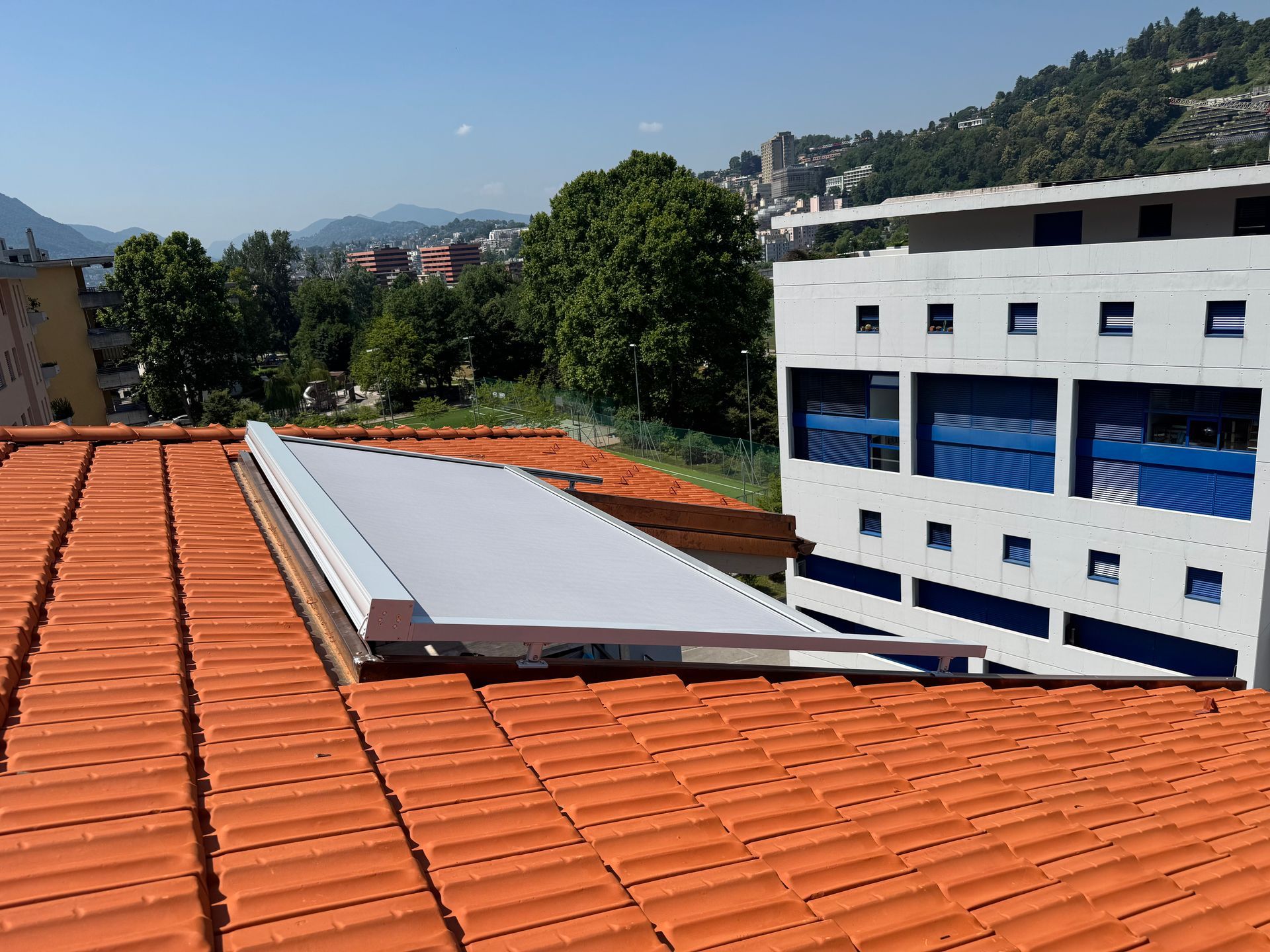 Una finestra per tetti bianca installata su un tetto inclinato di tegole in terracotta accanto a un moderno edificio a più piani.