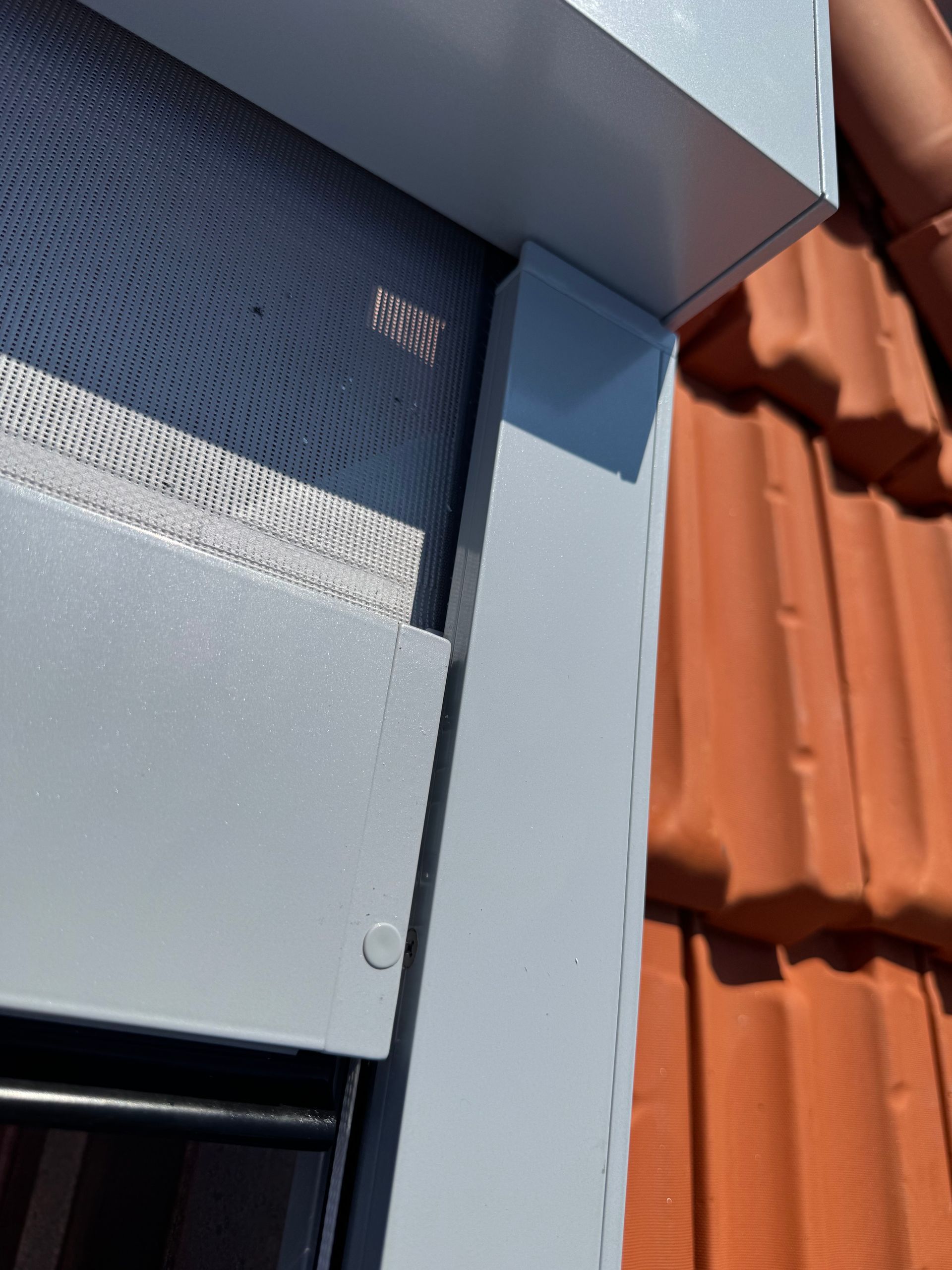 Primo piano di un telaio di finestra esterno grigio chiaro e del relativo binario per tenda parasole installati accanto a tegole di terracotta arancioni.
