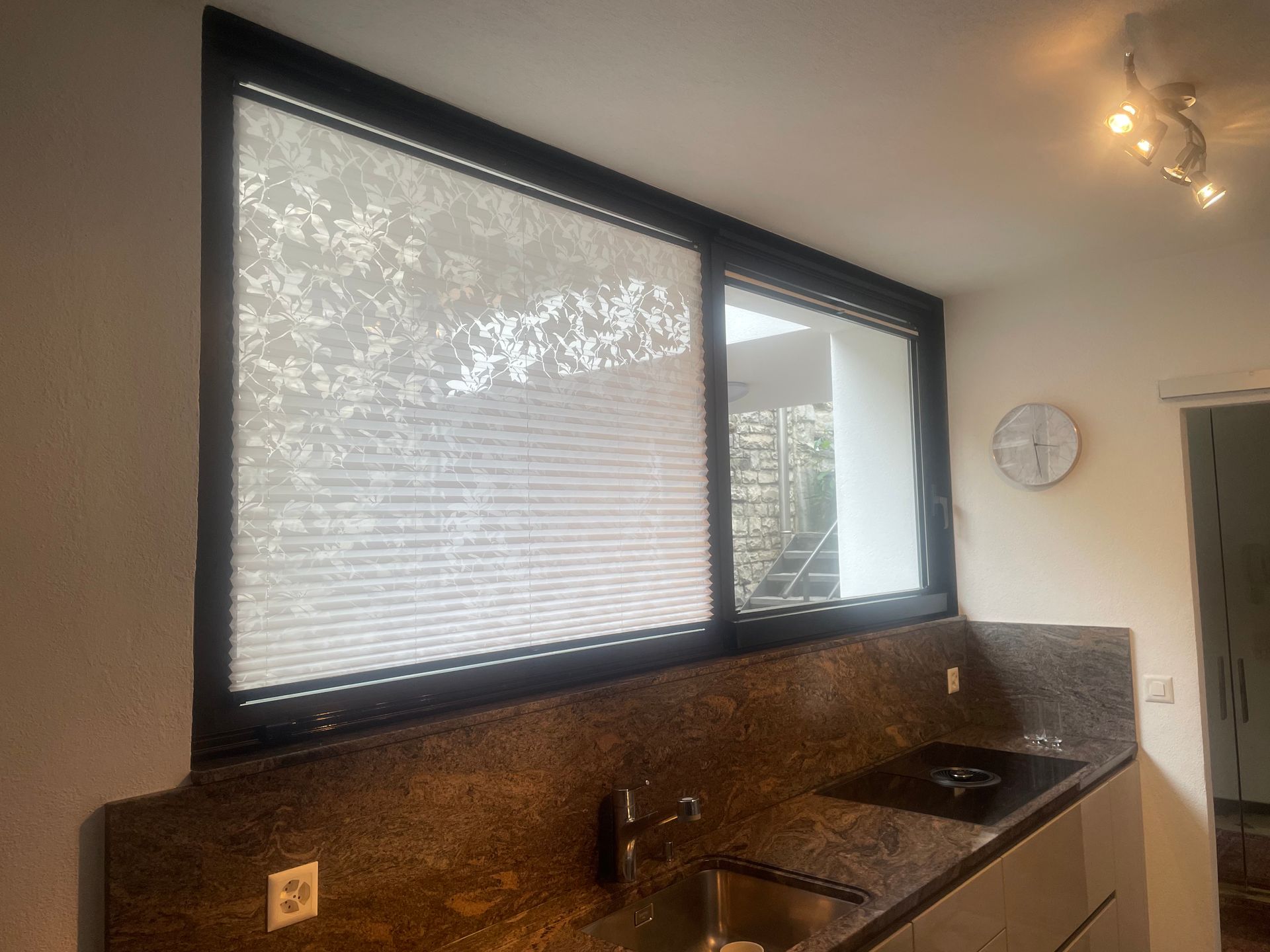 Una moderna zona lavello in cucina con ripiani in pietra scura, una finestra con tenda a pieghe e un lampadario a soffitto.