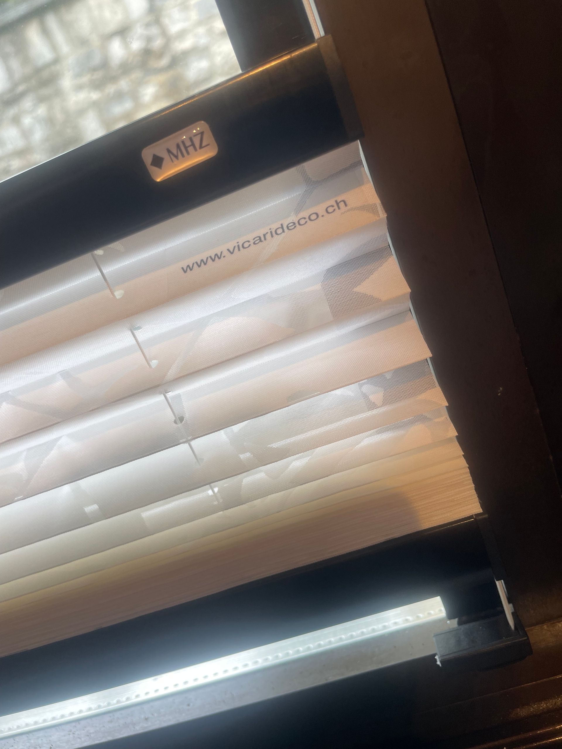 Primo piano di tende a pieghe bianche per finestre con una piccola etichetta nera che mostra il logo del marchio MHZ e il sito web.