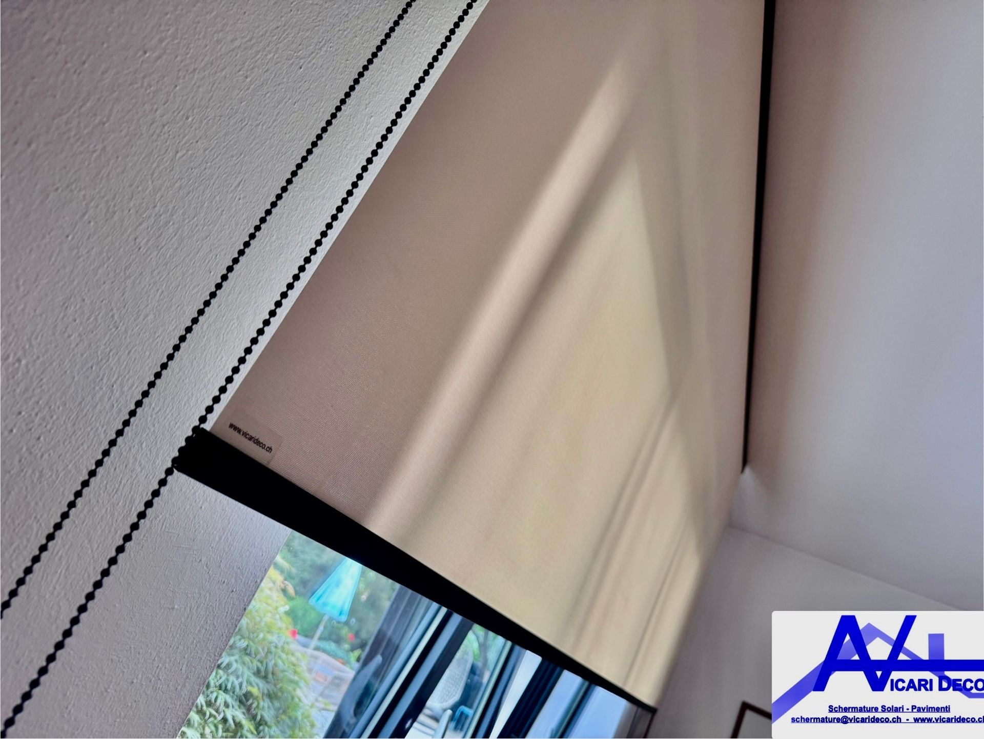 Una tenda a rullo beige parzialmente abbassata su una finestra, con una parete bianca dalla texture particolare e dettagli scuri nella cornice.