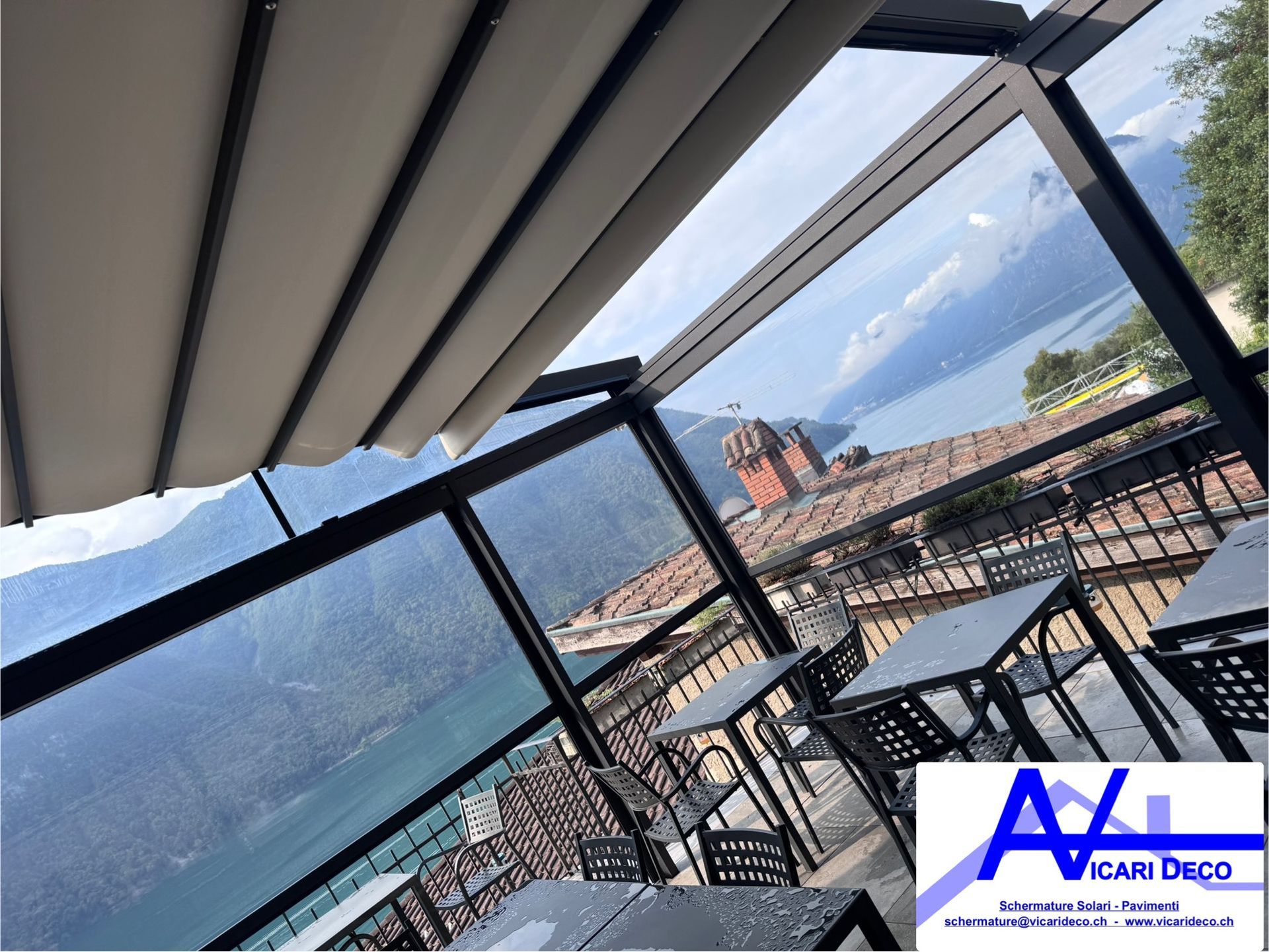 Una terrazza all'aperto con tavoli sotto un tetto retrattile, con vista su un lago, sulle montagne e su una torre di mattoni.