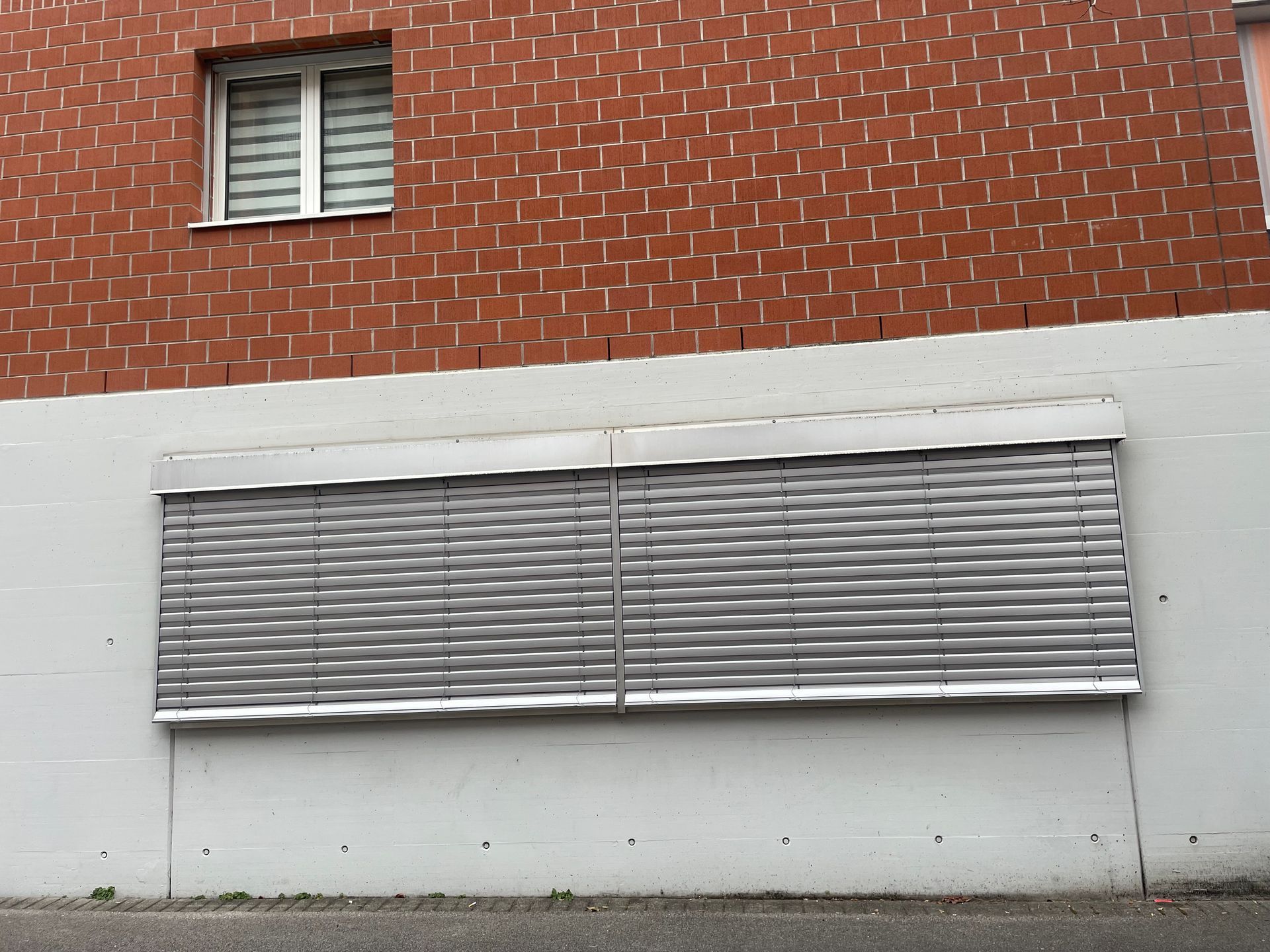 Una parete bianca con una grande persiana metallica chiusa, posizionata sotto un muro di mattoni con una singola piccola finestra.
