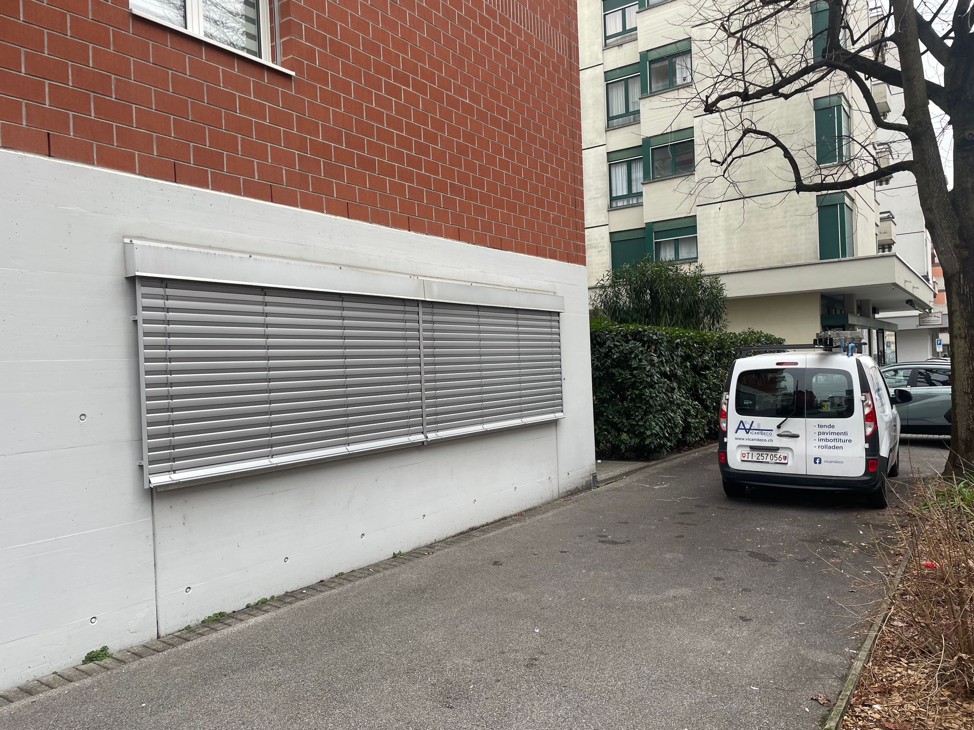 Un furgone bianco parcheggiato accanto a un edificio con una grande persiana a lamelle su una parete bianca e rossa.