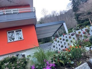 Una casa arancione brillante con una terrazza, un balcone grigio e un muro di contenimento a gradoni ricoperto di piante e fiori.