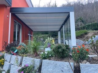 Una moderna copertura per patio esterno con una struttura scura fissata a una casetta arancione brillante, posizionata sopra un muro di pietra da giardino.