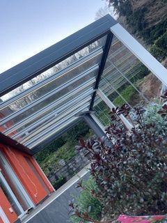 Una pergola in metallo scuro con tetto in vetro, annessa a una casa dalle pareti arancioni, vista da un giardino.
