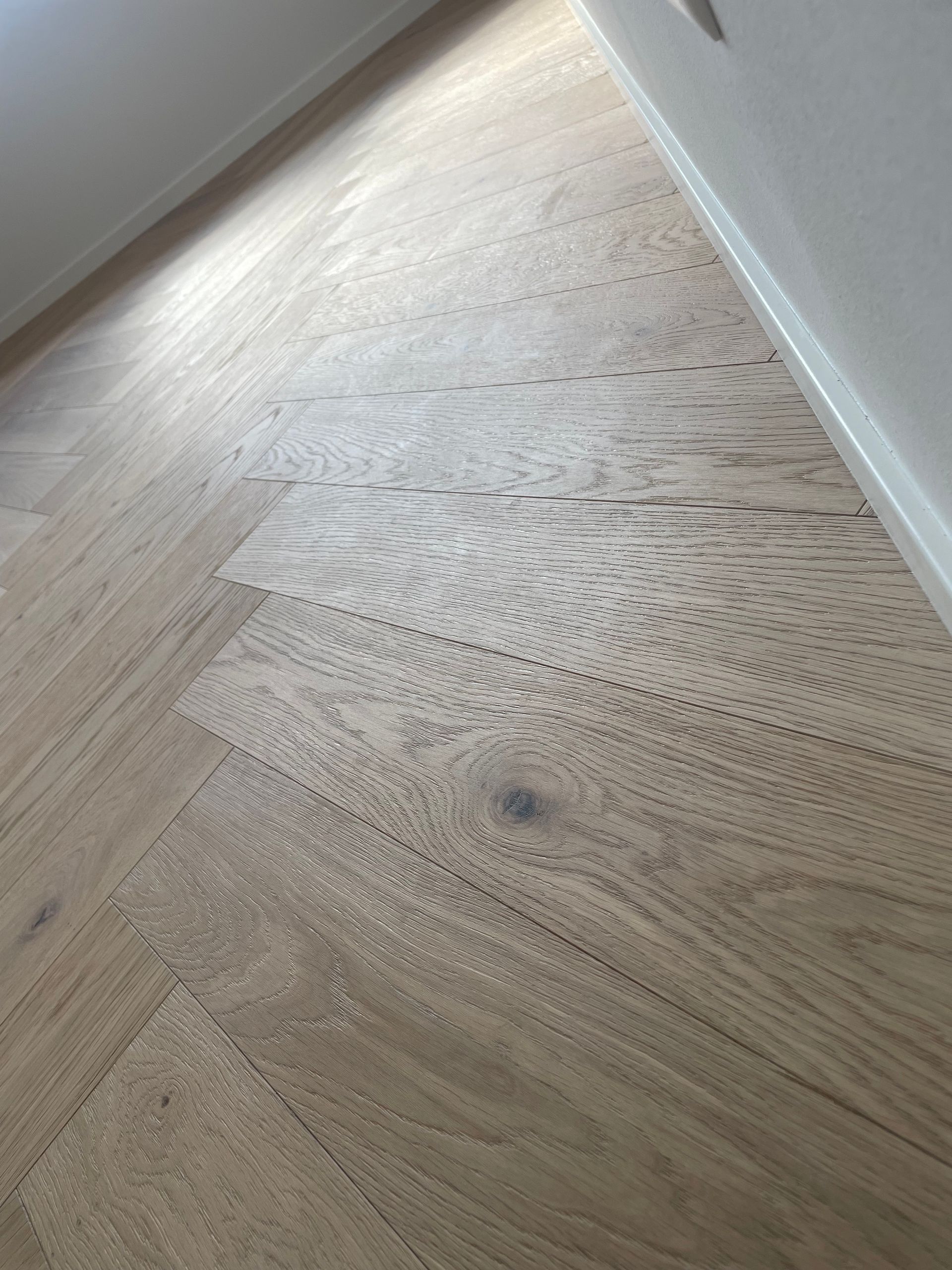 Un primo piano, ripreso dall'alto, di un pavimento in legno chiaro con motivo a spina di pesce che incontra un battiscopa bianco.