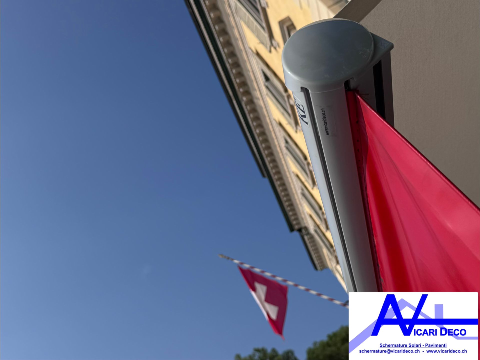 Una tenda da sole rossa fissata a un edificio, con una bandiera svizzera che sventola sullo sfondo, contro un cielo azzurro e limpido.
