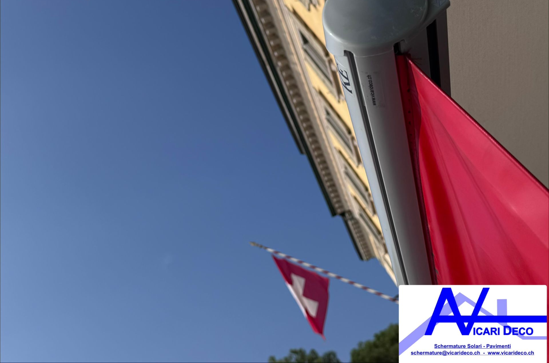 Una tenda di tessuto rosso si estende dalla facciata di un edificio stagliandosi contro un cielo azzurro brillante, con una bandiera svizzera che sventola in lontananza.