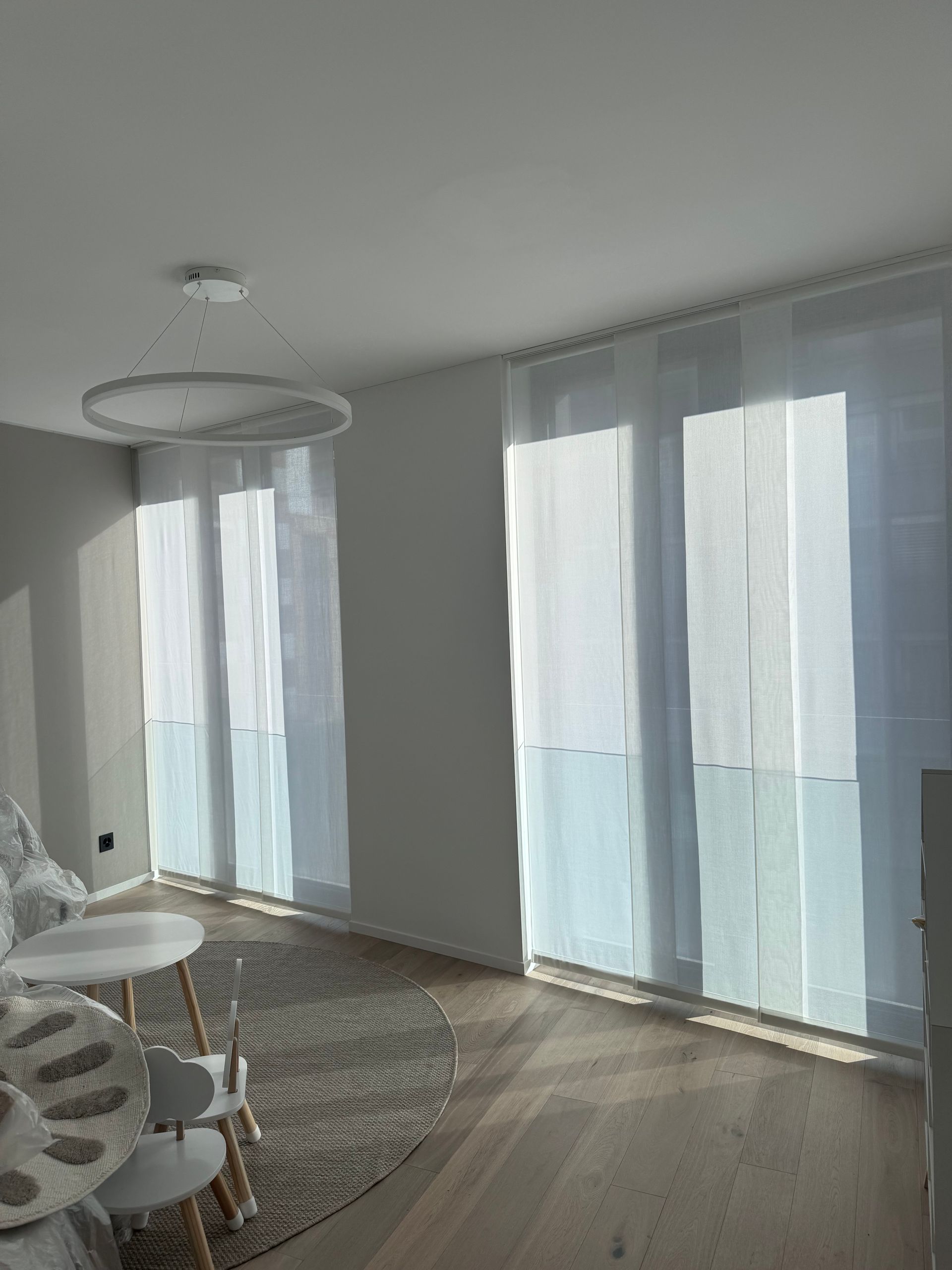 Soggiorno moderno con tappeto rotondo, piccolo set da pranzo, lampada a sospensione circolare e tende trasparenti alle finestre.