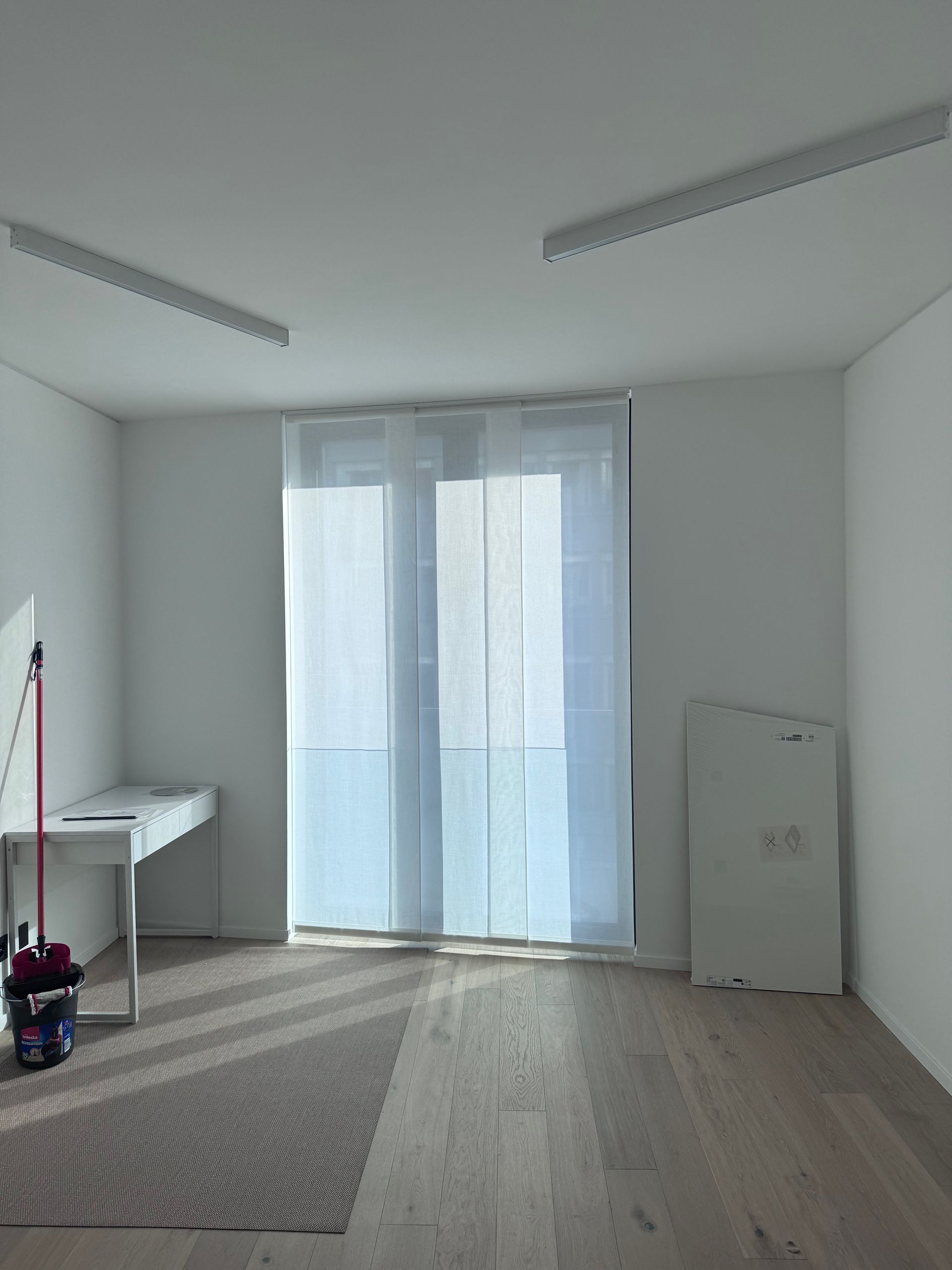 Una stanza moderna e minimalista con pavimento in legno chiaro, pareti bianche, una scrivania sulla sinistra, tende trasparenti e illuminazione a incasso.