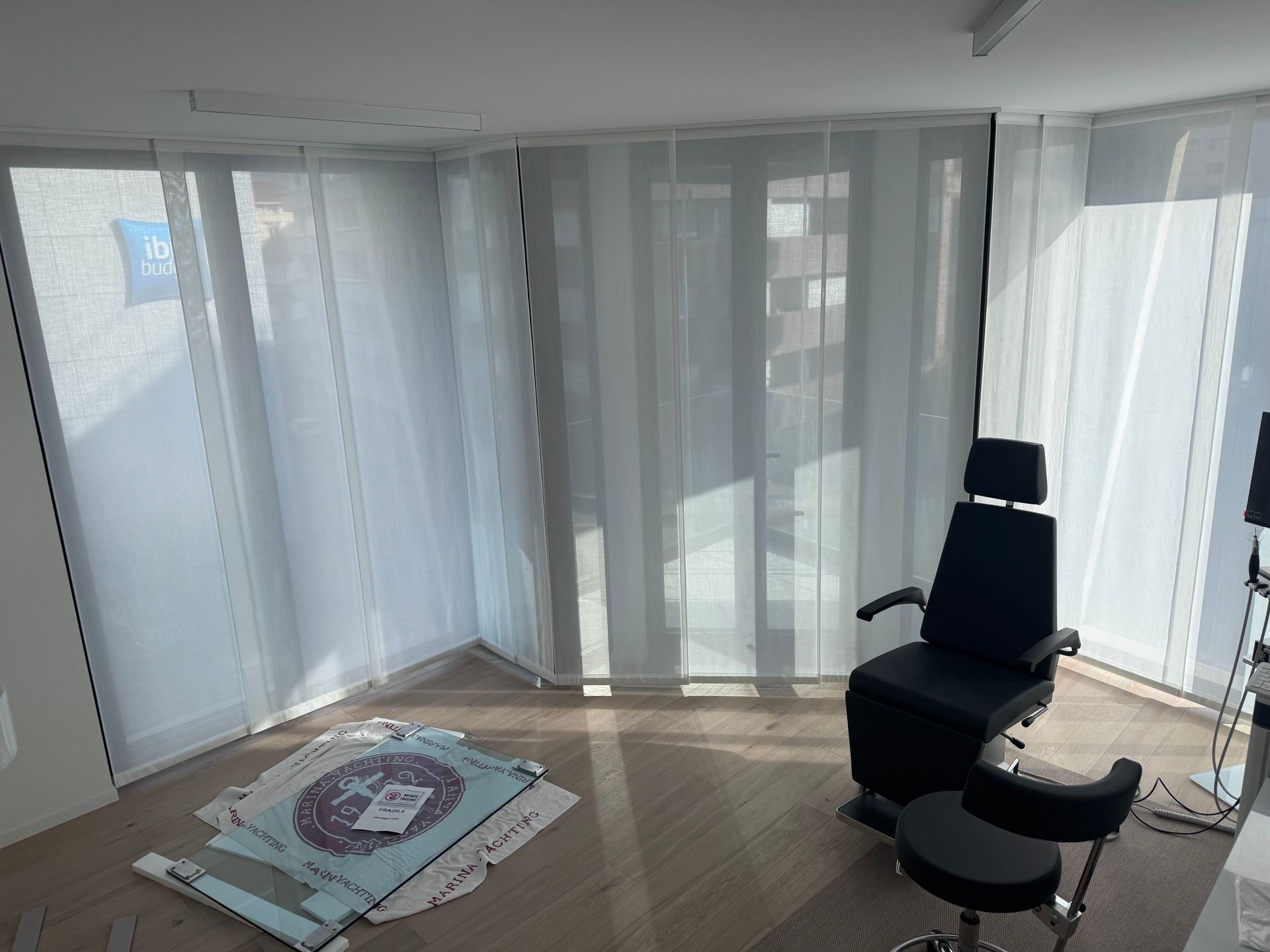 Una stanza da ambulatorio minimalista con poltrona per trattamenti, sgabello e tende leggere che coprono le finestre a tutta altezza.