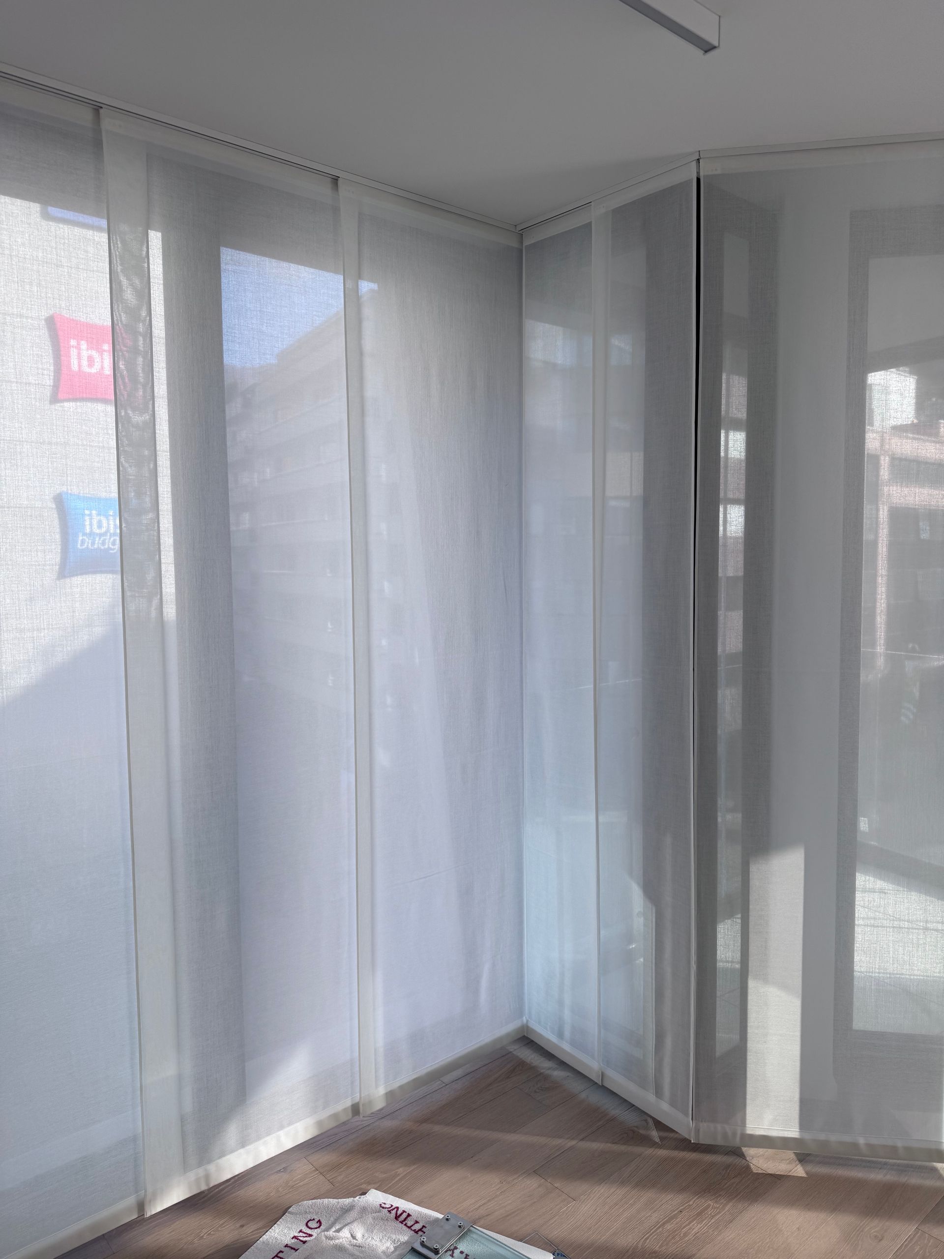 Delle tende a pannello bianche e trasparenti, appese a un binario sopra una finestra ad angolo, filtrano la luce solare intensa.