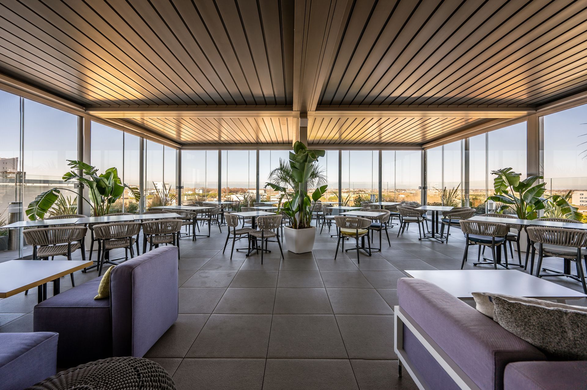 Ristorante moderno sul tetto con tavoli all'aperto, divani viola e grandi piante in vaso sotto un soffitto a doghe.