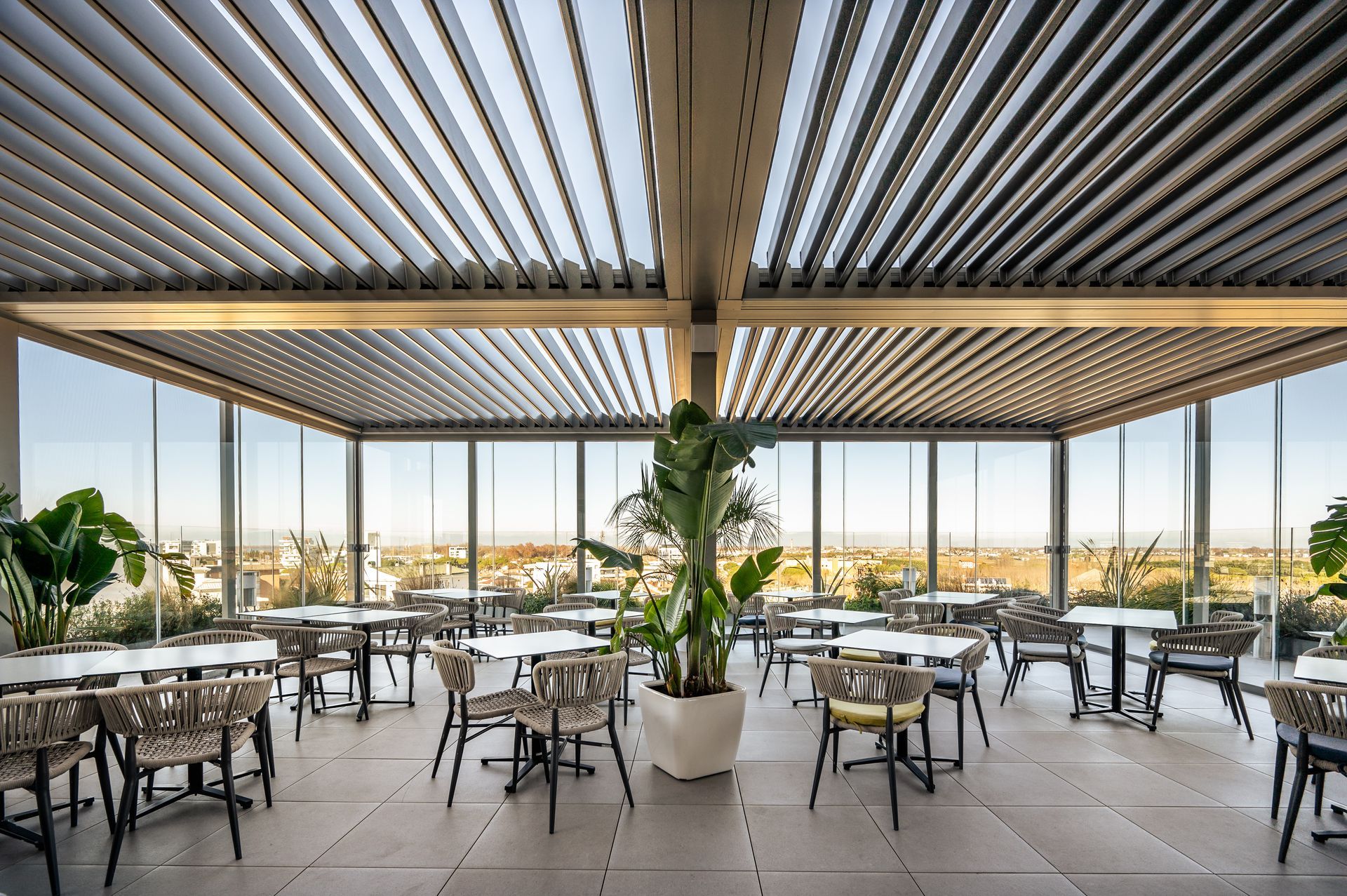 Un patio ristorante all'aperto con soffitto a lamelle metalliche, pareti in vetro, tavoli da pranzo, sedie e piante in vaso.