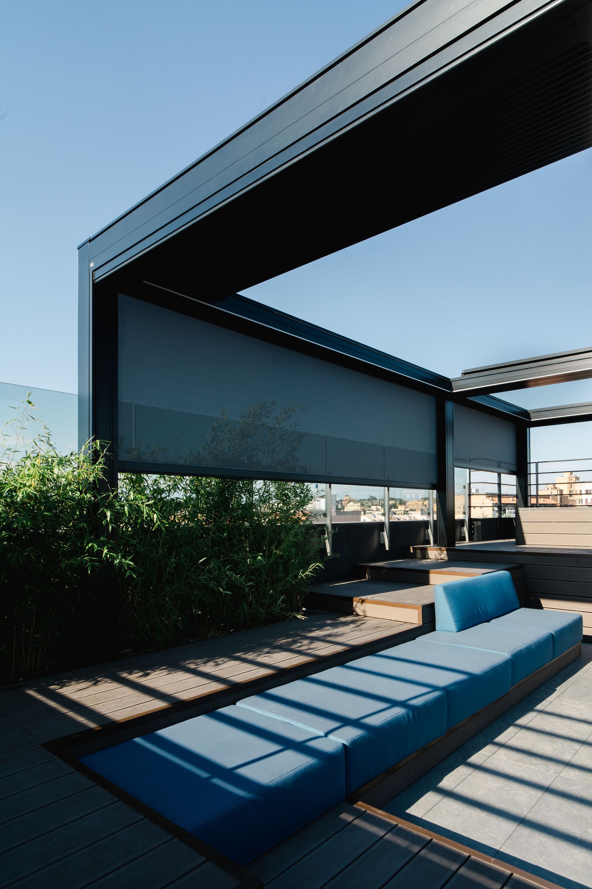 Una panchina blu è posizionata su una terrazza di legno soleggiata, sotto un moderno pergolato in metallo nero con tende parasole retrattili in tessuto.